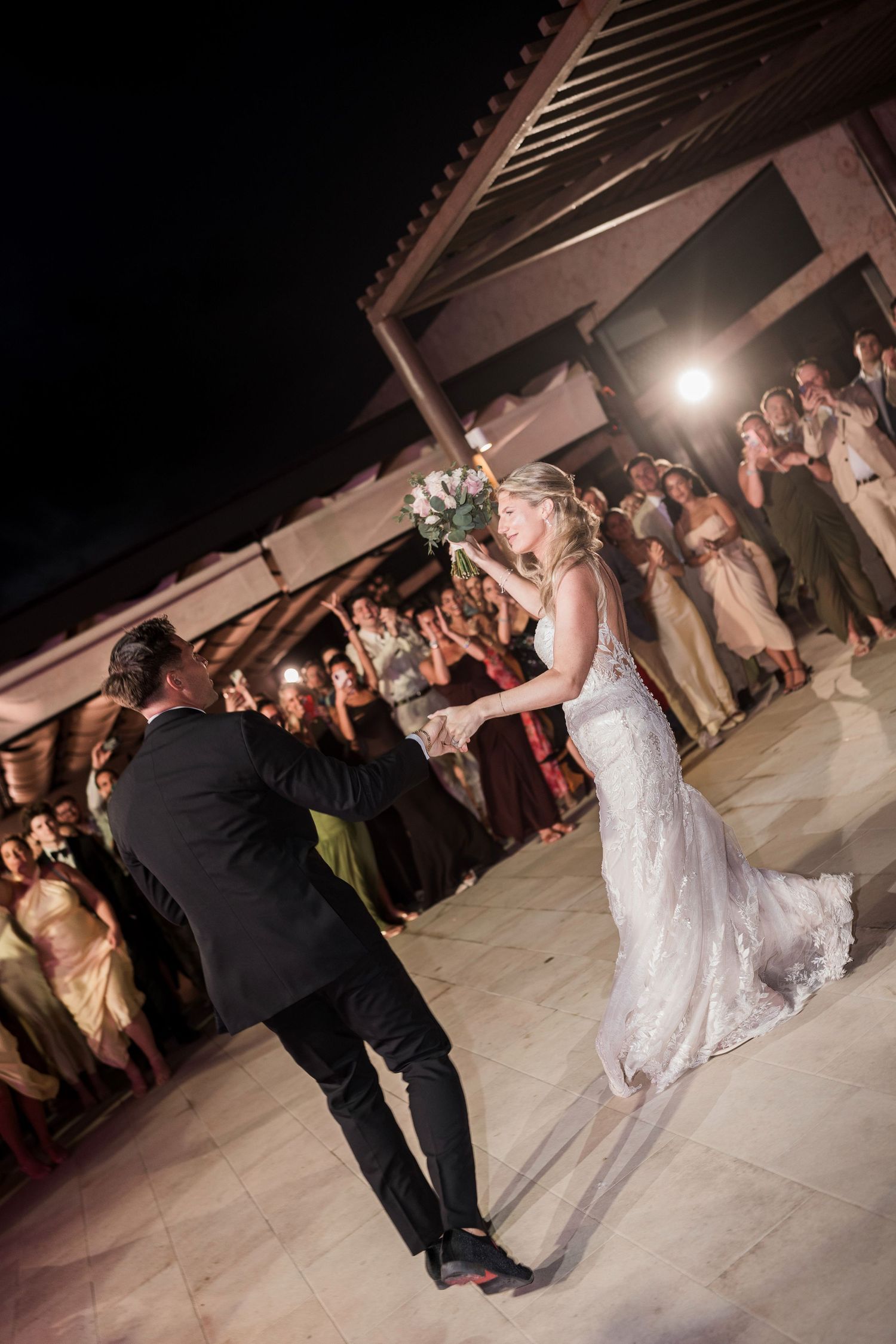 Bride and groom share their first dance at an outdoor wedding reception at night, surrounded by cheering guests.