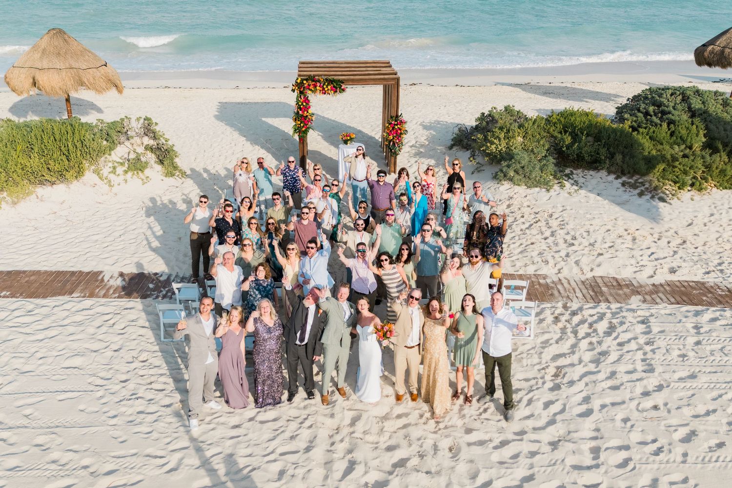 Large wedding group photo on a sandy beach with a wooden arch decorated with tropical flowers and turquoise ocean backdrop.