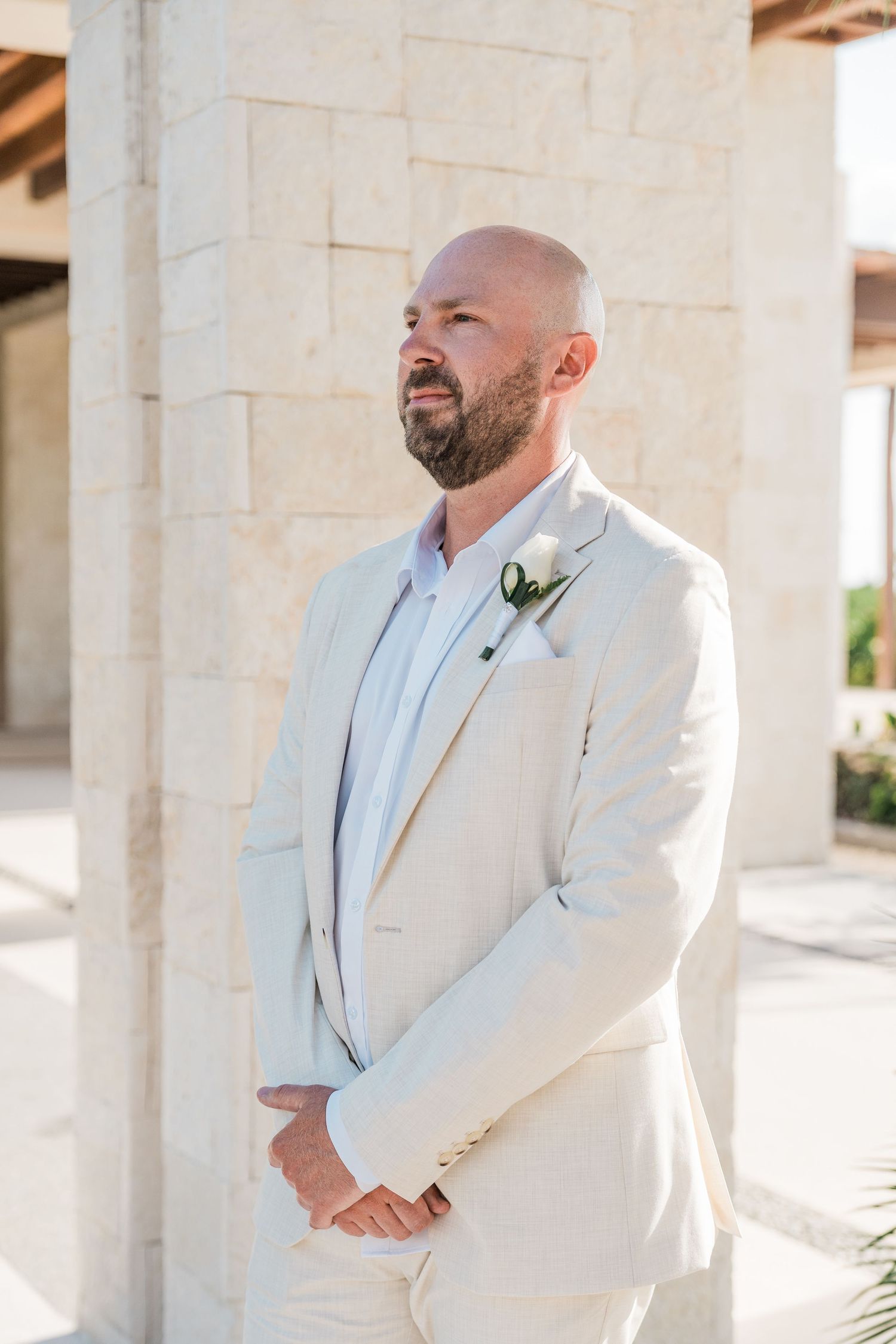 Bald groom with beard in cream linen suit and floral boutonniere stands outdoors by stone columns.