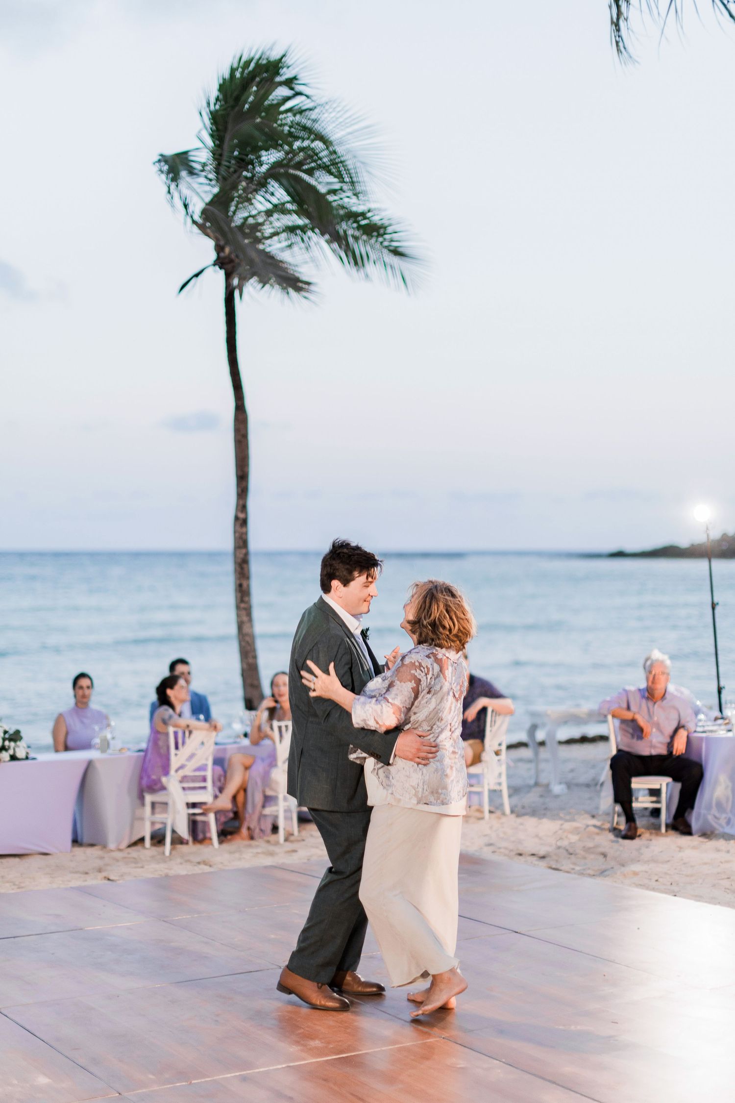 Couple shares a romantic mother-son dance on a beachside outdoor dance floor at a tropical wedding reception.