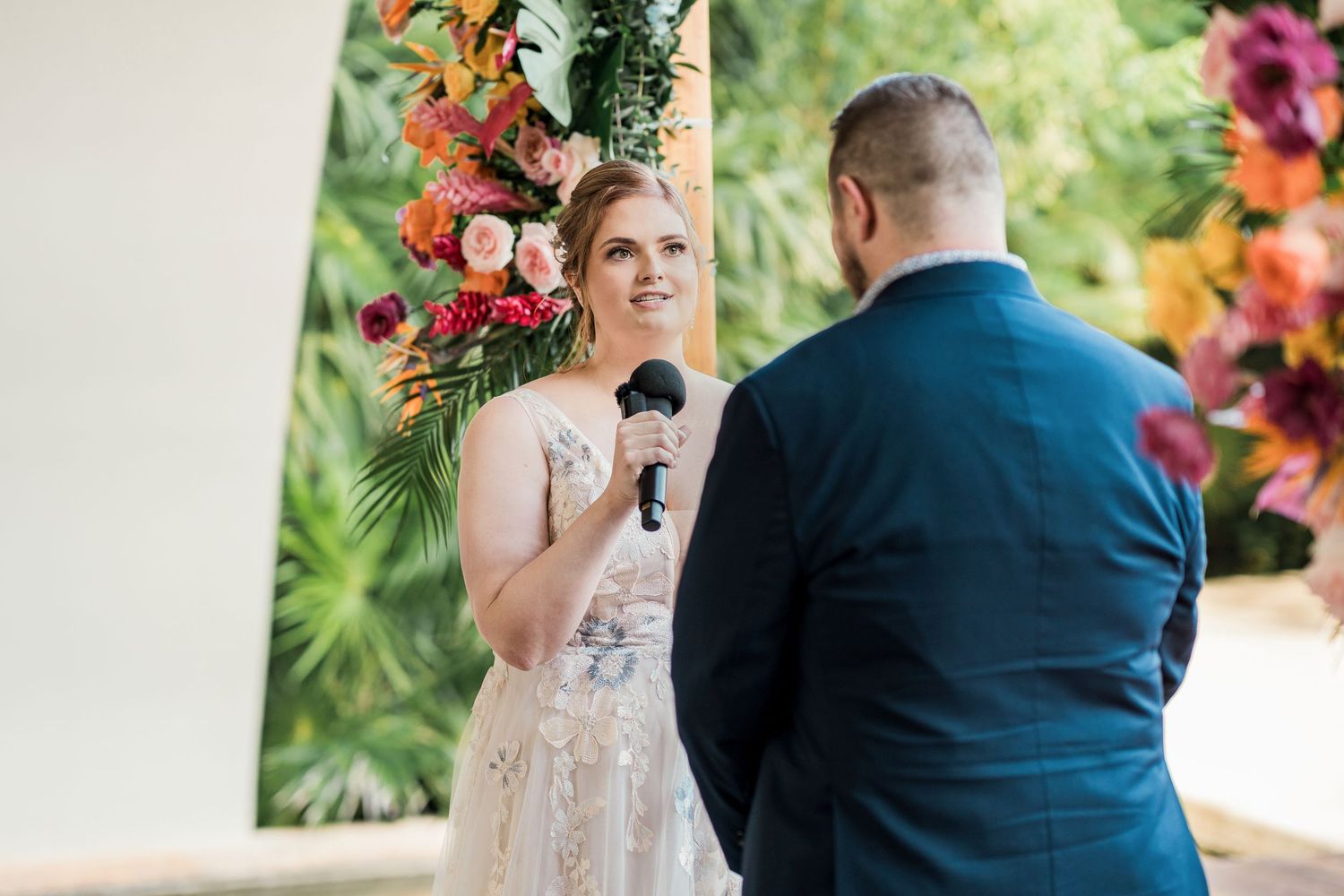 Secrets Akumal Wedding - Bride smiling while reciting vows at outdoor wedding ceremony with colorful tropical floral arch backdrop.