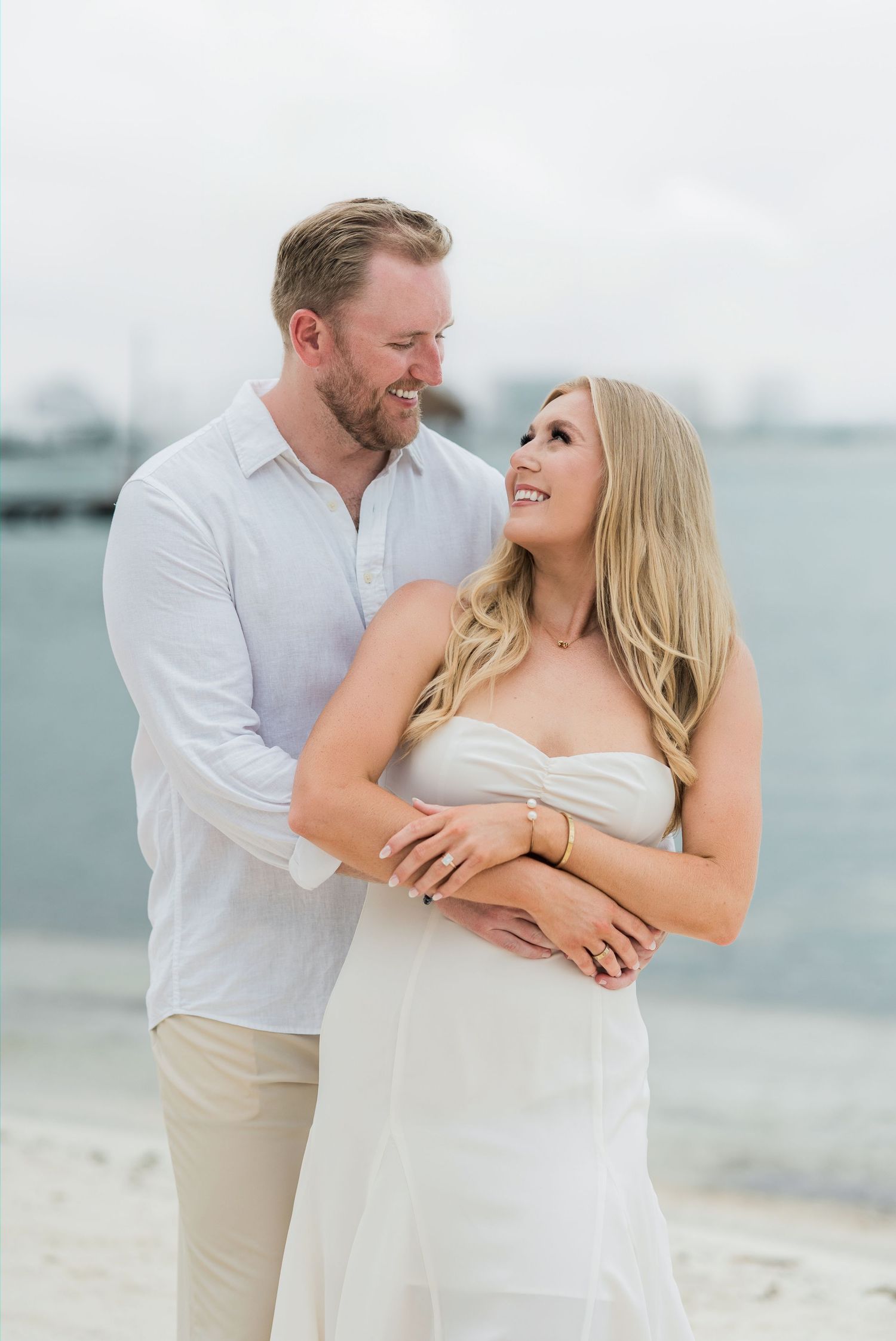 Cancun Engagement Photoshoot - Happy couple embracing on a sandy beach, wearing white outfits, smiling at each other