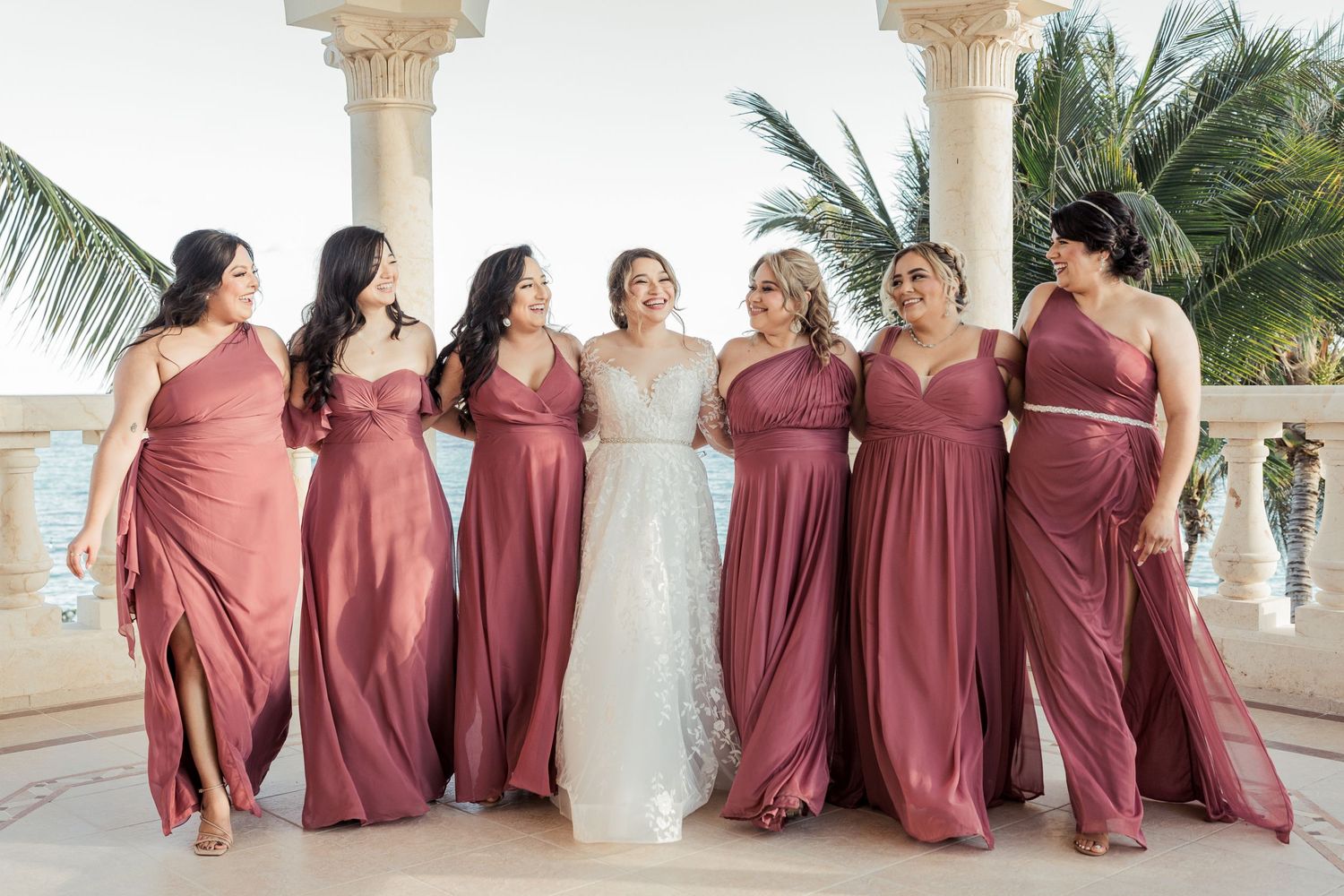 Six bridesmaids in flowing dusty rose gowns walking with bride in white lace dress at elegant beachfront venue.