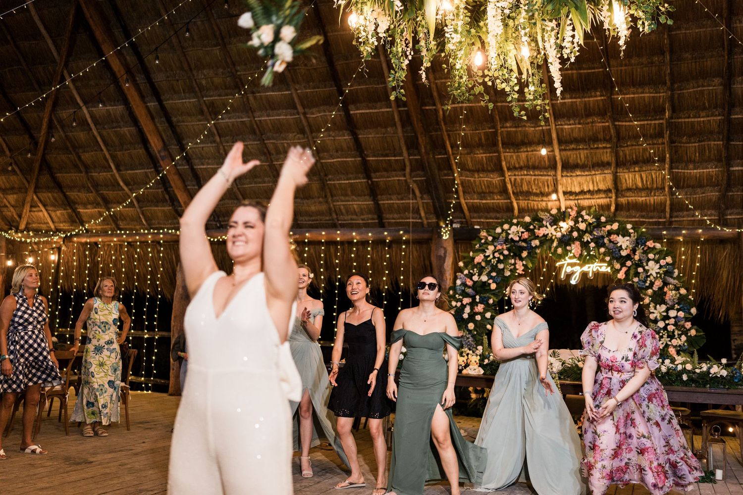 Bride tossing bouquet to bridesmaids in sage green dresses at rustic thatched roof wedding venue.