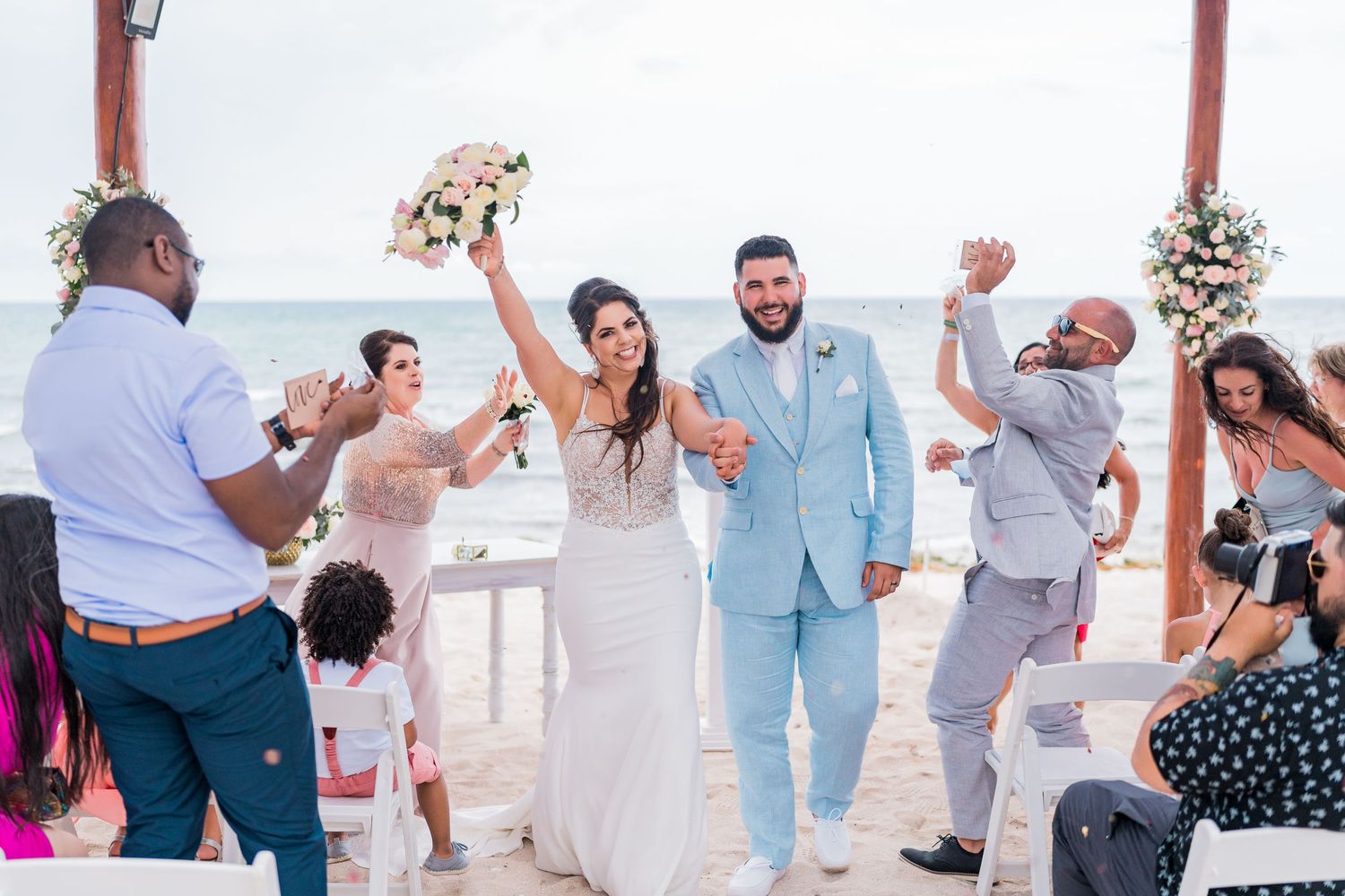 Joyful newlyweds celebrate their recessional on the beach surrounded by cheering wedding guests.