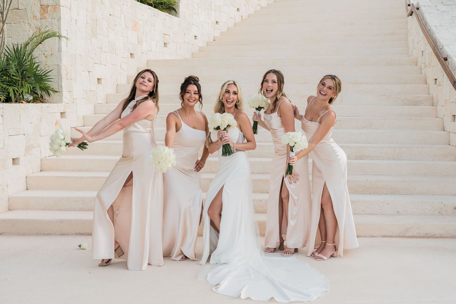 Bride and bridesmaids in blush gowns laugh and pose playfully together on elegant white stone stairs.