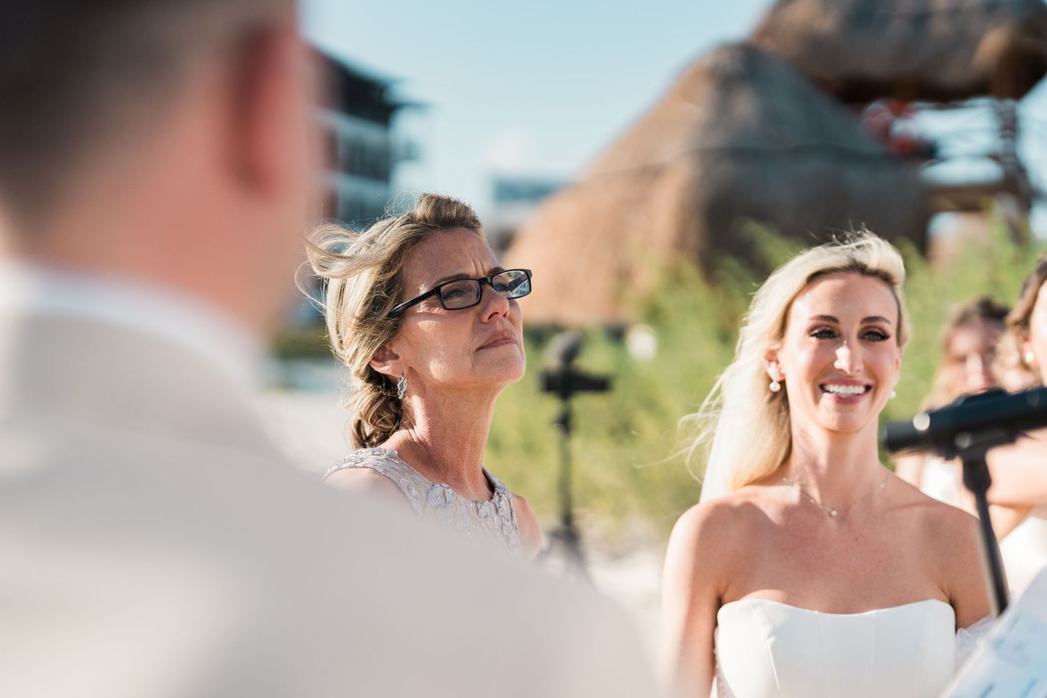 Smiling bride in white gown stands at beach altar with emotional mother beside her during outdoor ceremony.