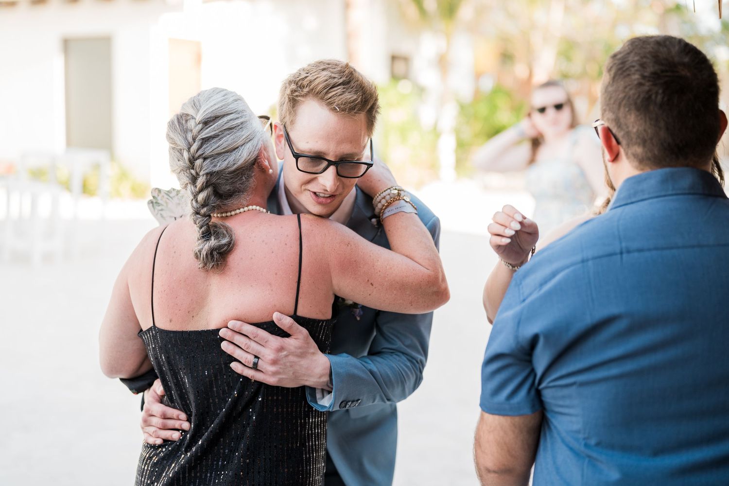 Groom in blue suit shares heartfelt hug with older woman at tropical wedding celebration.