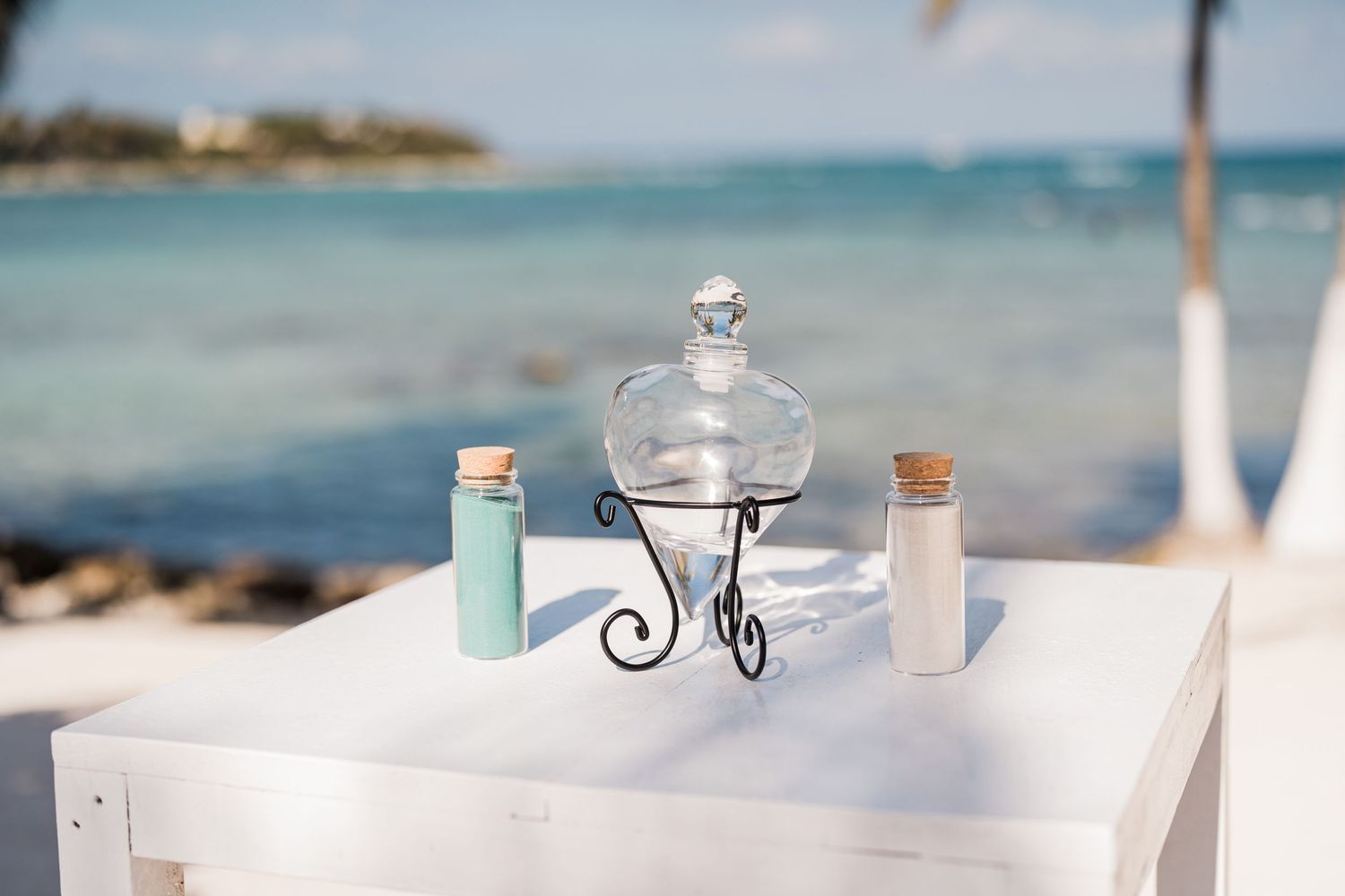 Sand ceremony setup with colorful sand bottles and glass vase on white table by the ocean.