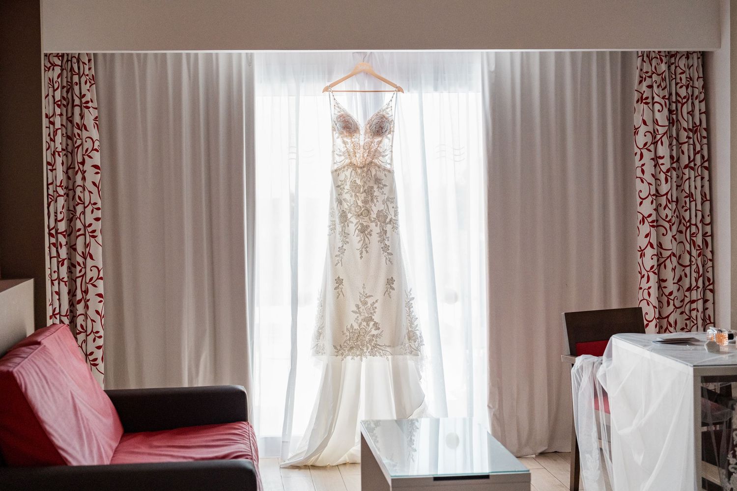 Beautiful beaded lace wedding dress hanging on a wooden hanger in front of sheer white curtains.