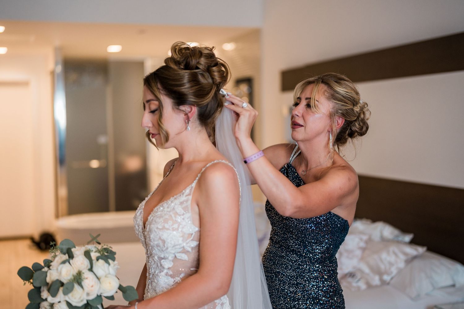 Woman helping bride attach veil to elegant updo in hotel room.