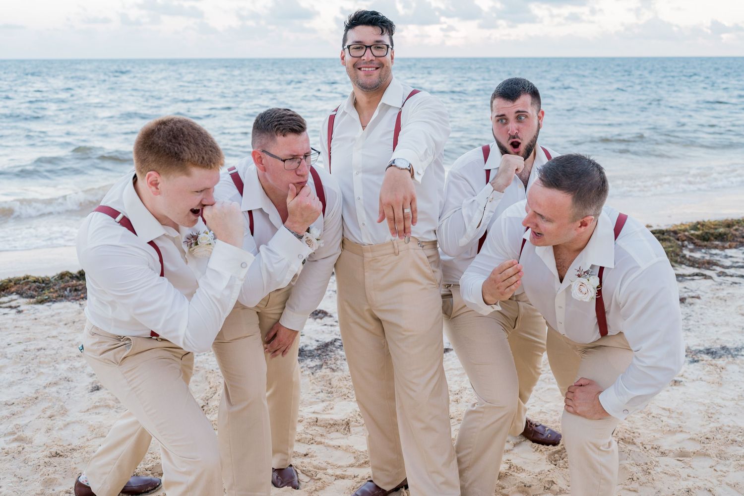 Groom and groomsmen in white shirts with burgundy suspenders posing playfully on beach.