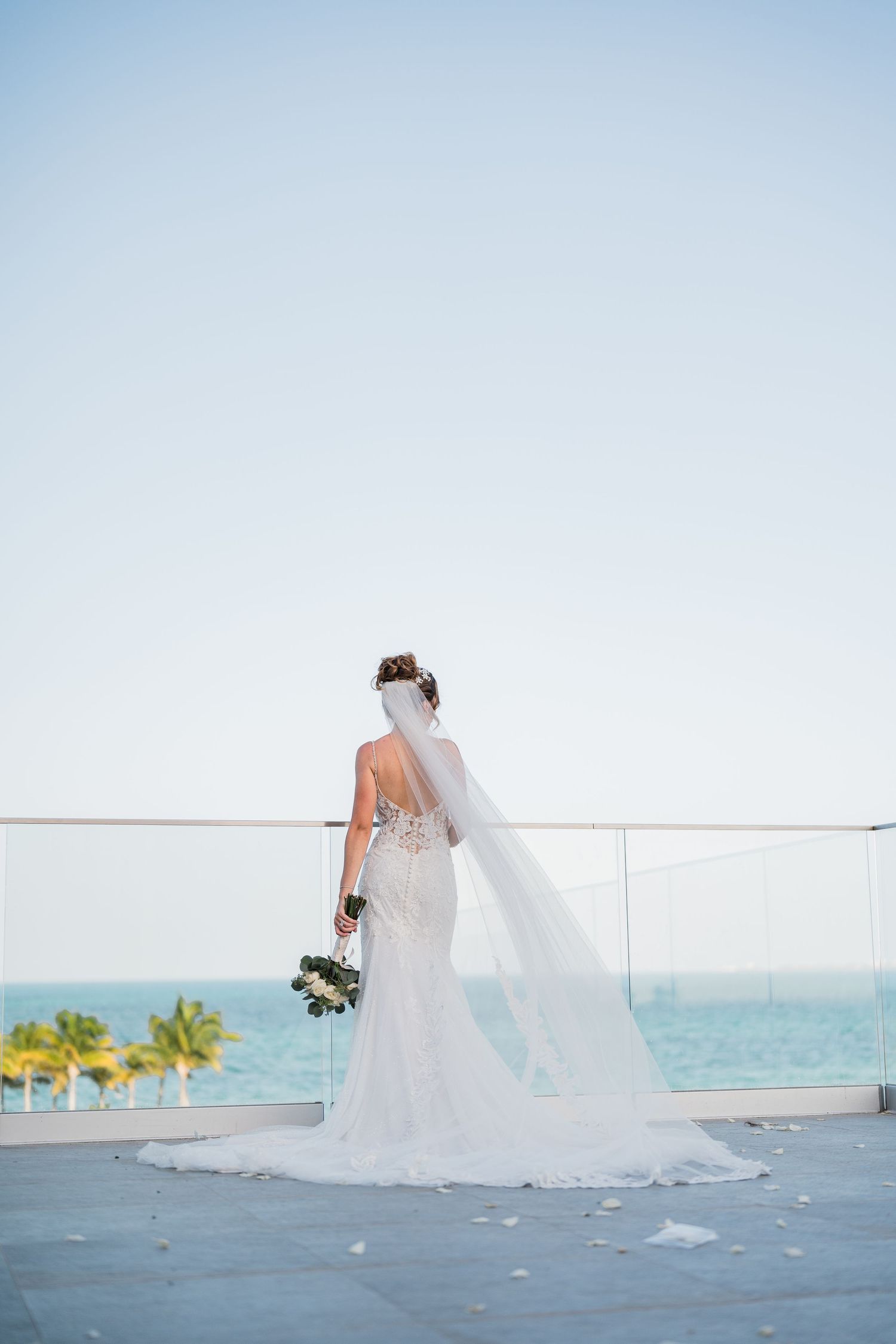 Bride in fitted lace gown with long veil stands gazing at turquoise ocean view.