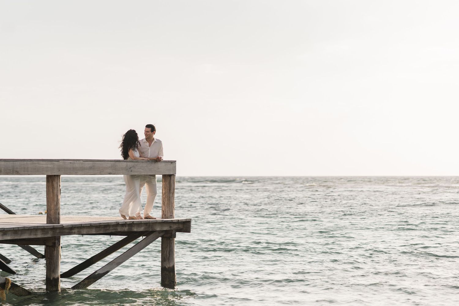 Couple embracing on a wooden pier overlooking a calm ocean under a bright sky.