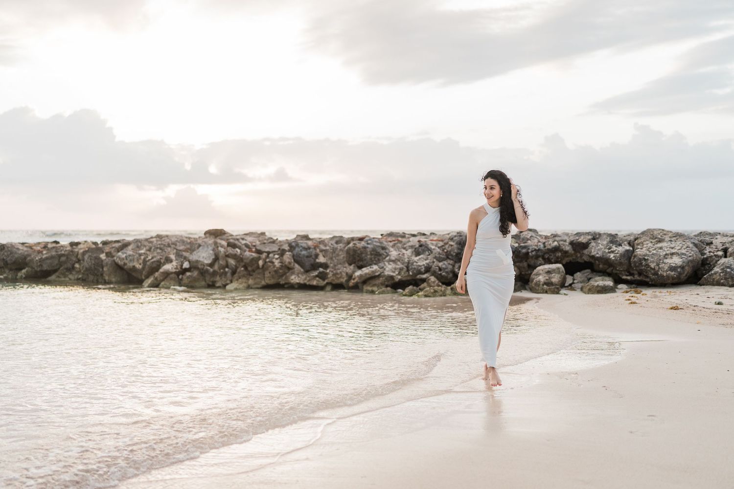 A woman in a white dress walks along a sandy beach near rocky jetty at sunset.
