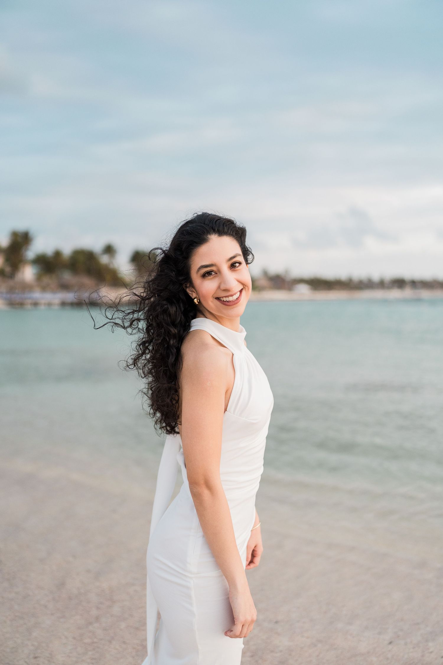 A smiling woman in a white sleeveless dress stands on a tropical beach with turquoise water in the background.