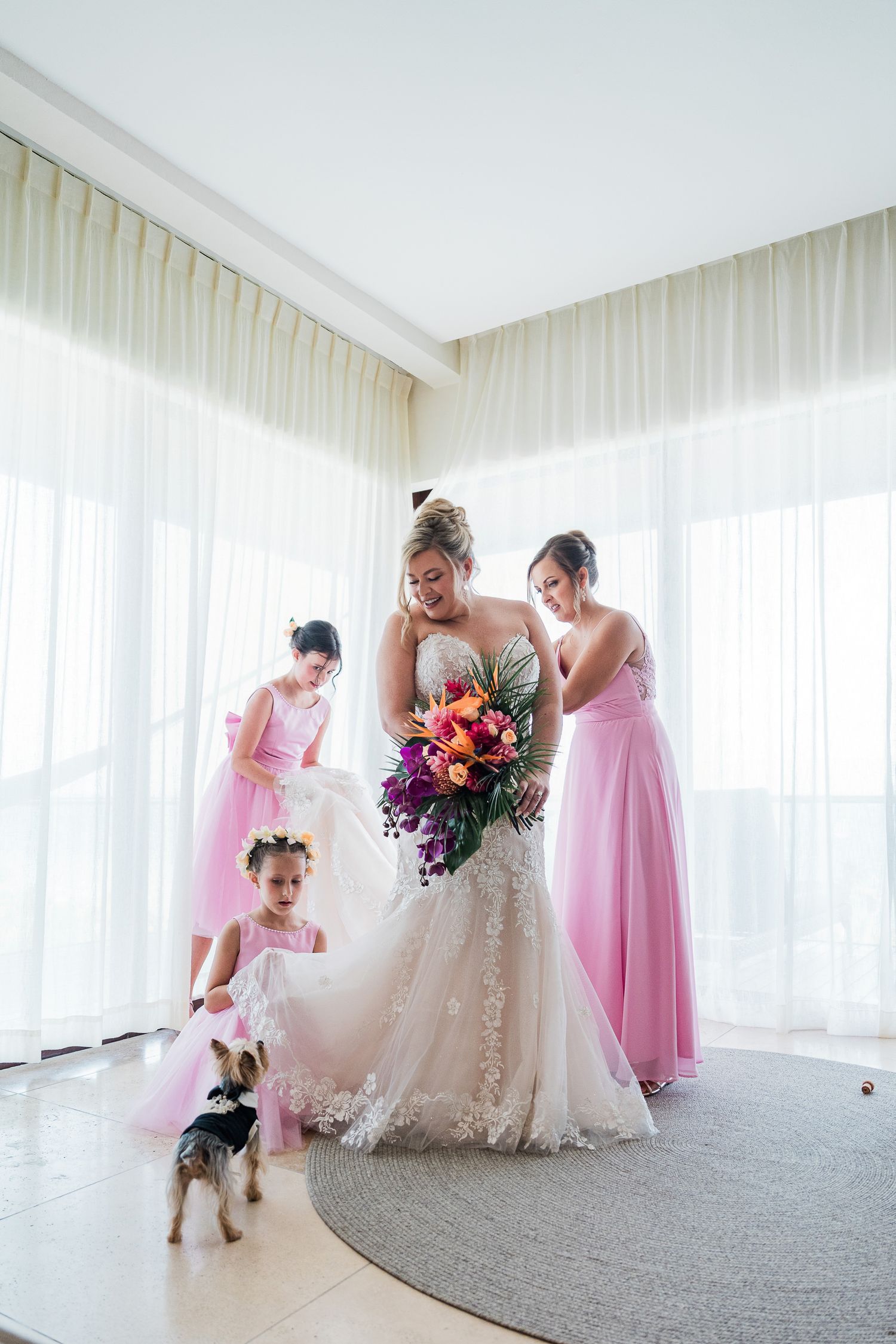 Bride in lace gown holds colorful bouquet with bridesmaids in pink dresses and a small dog nearby in a bright room.