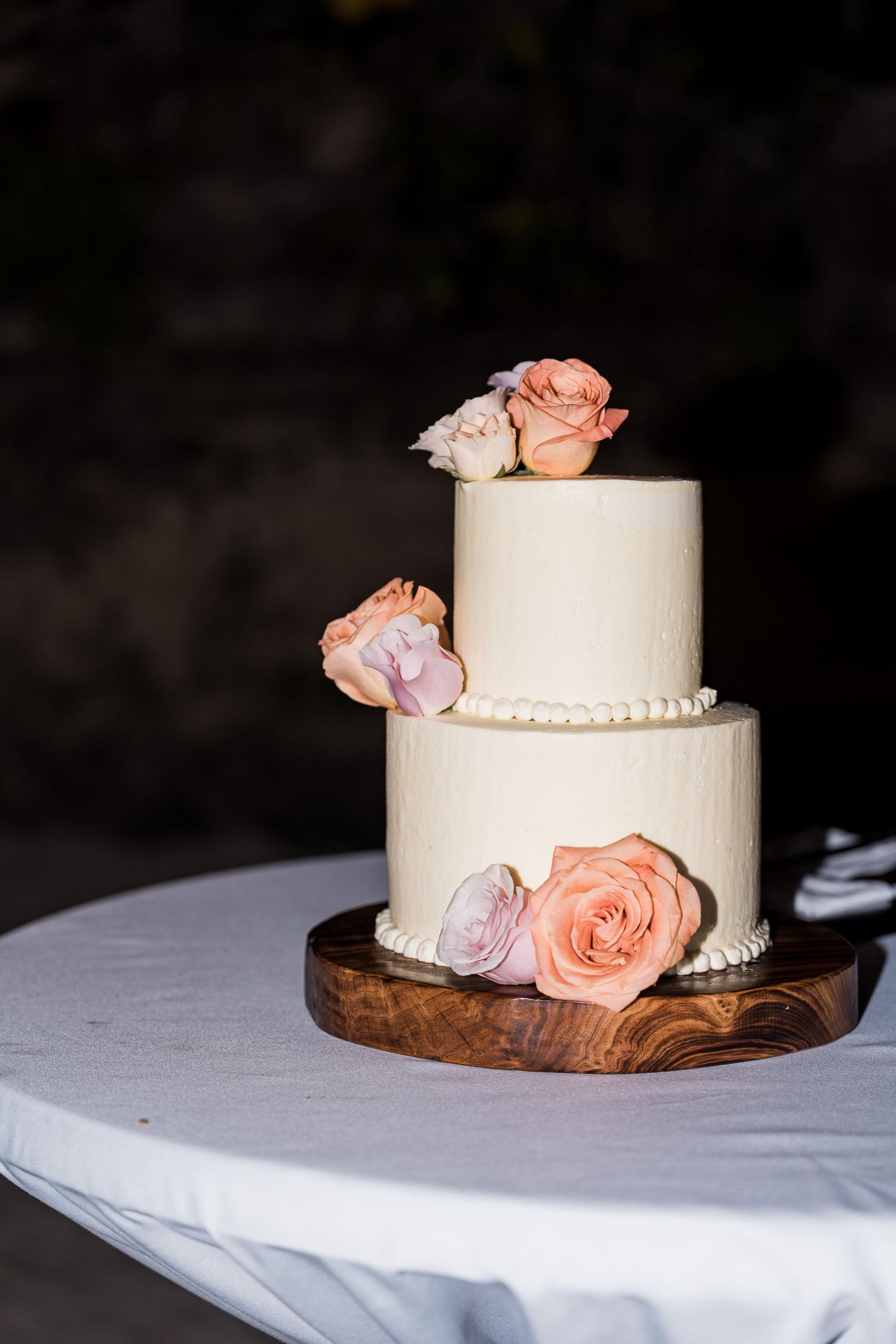 Hotel Xcaret México Two-tier white wedding cake decorated with peach and pink roses on a wooden board, displayed on a white tablecloth.
