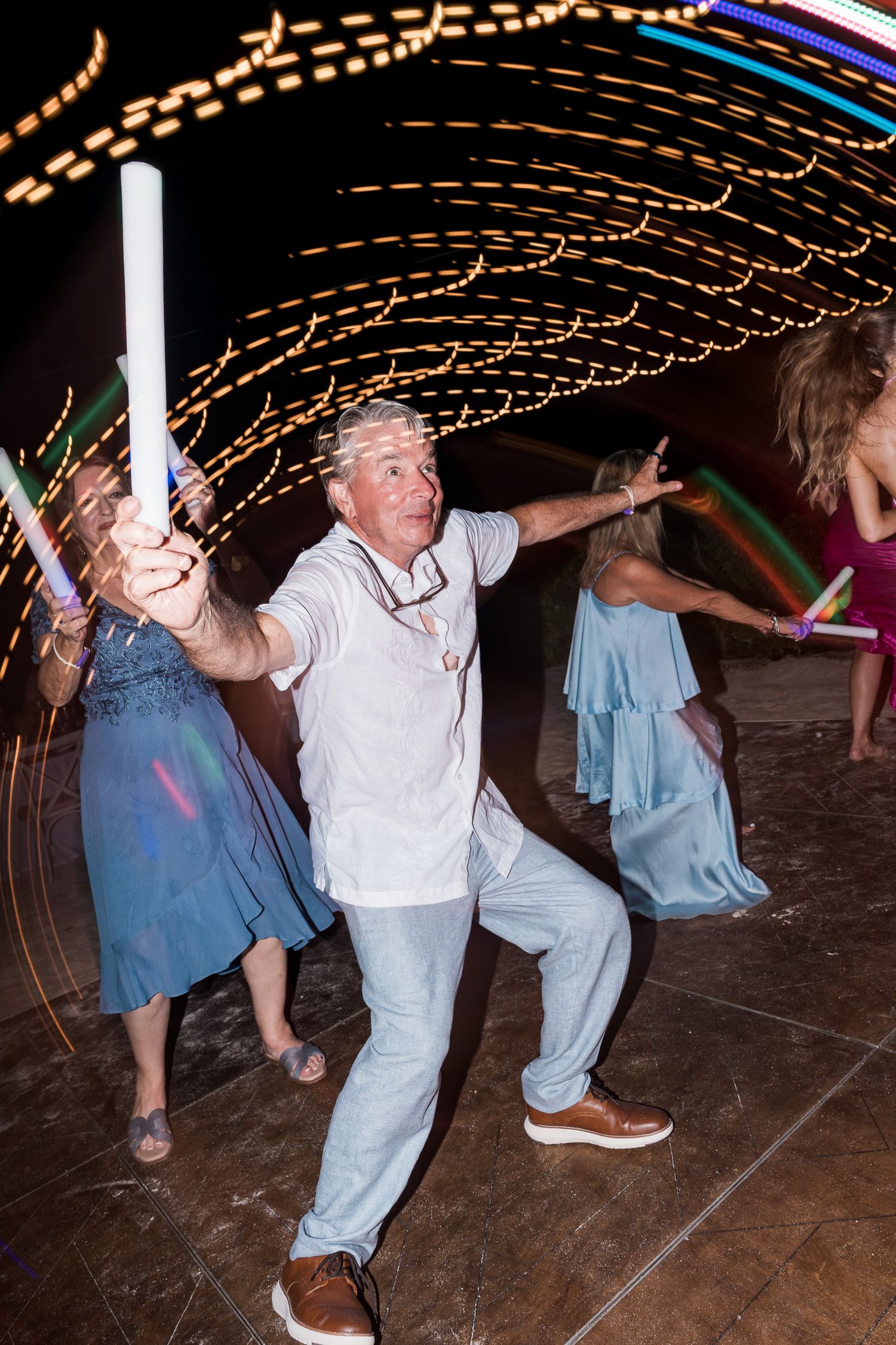 Man dancing energetically with glow stick at night party, colorful light trails swirling in background.