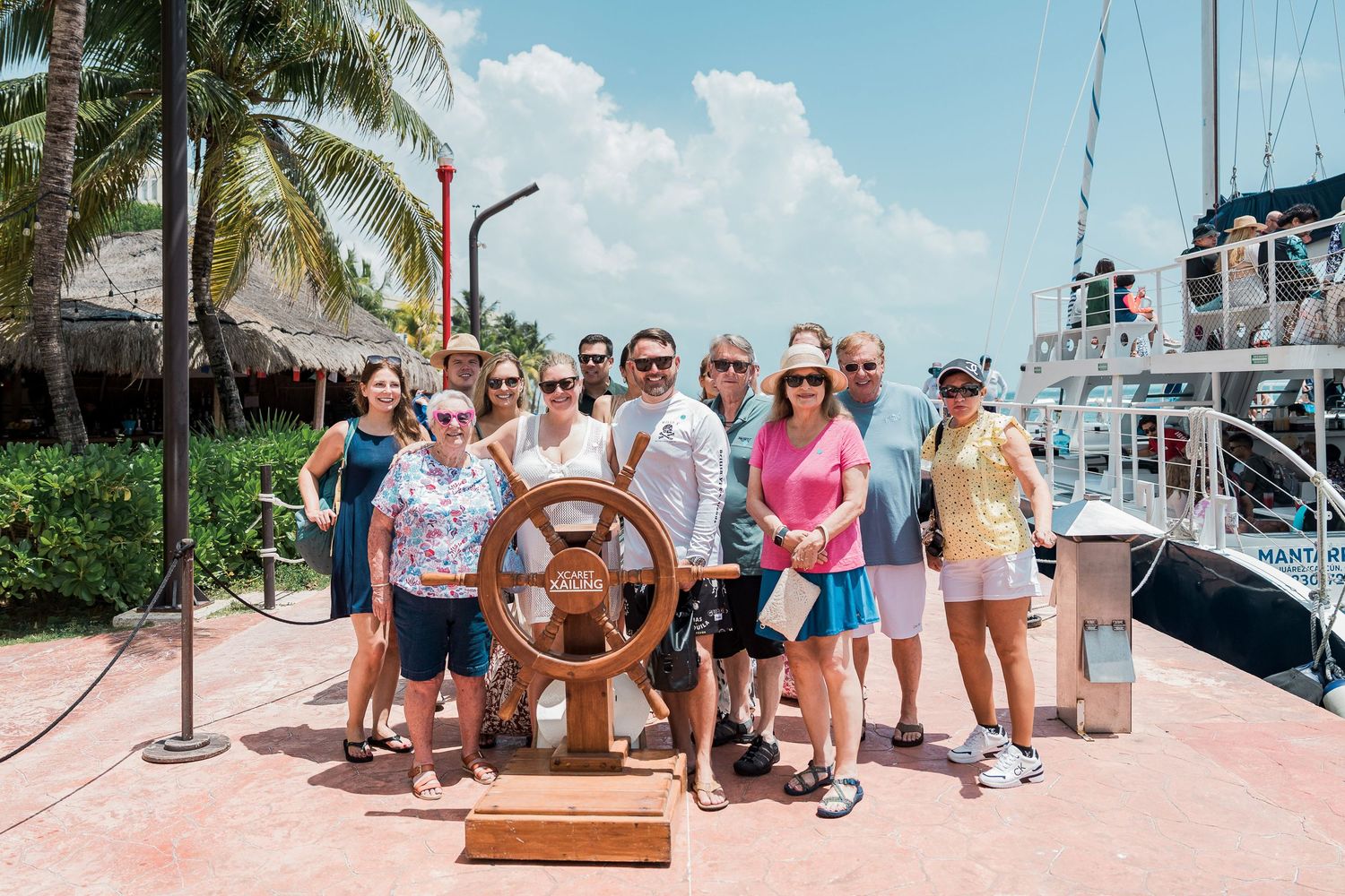 Riviera Maya Multi Day wedding - Group posing at a ship steering wheel on a sunny marina dock with sailboats and palm trees in background.