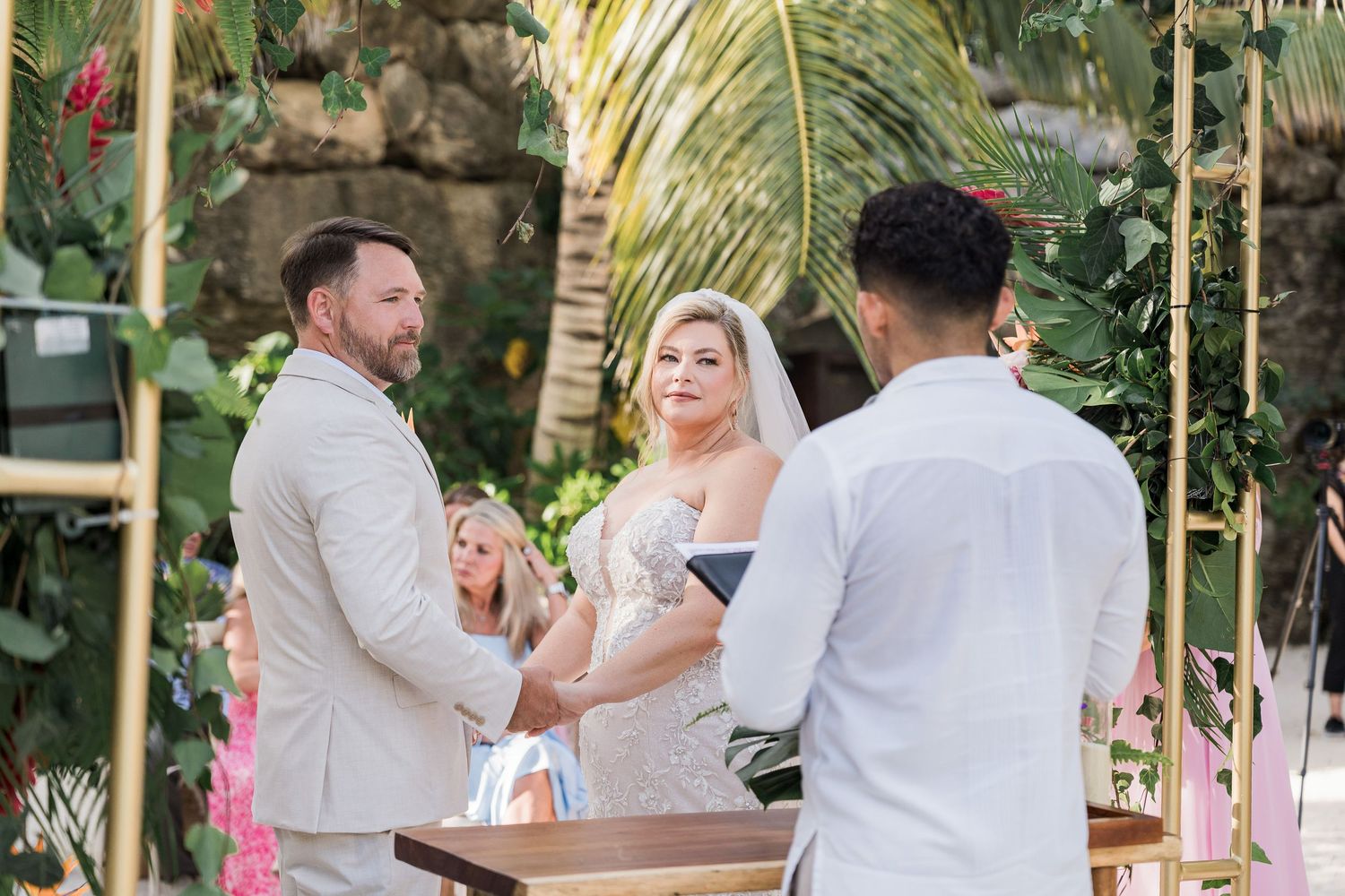Bride and groom exchange vows at tropical outdoor wedding ceremony with lush greenery and pink floral decorations.