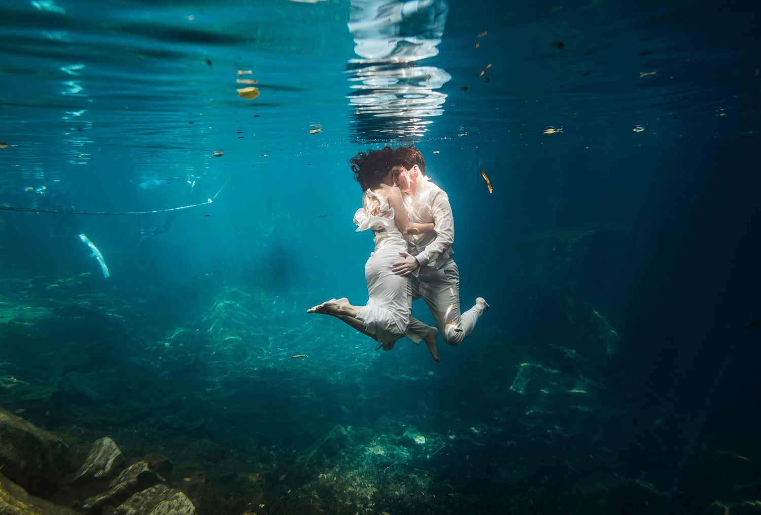 Couple in white attire sharing a romantic underwater embrace in a crystal clear turquoise ocean.