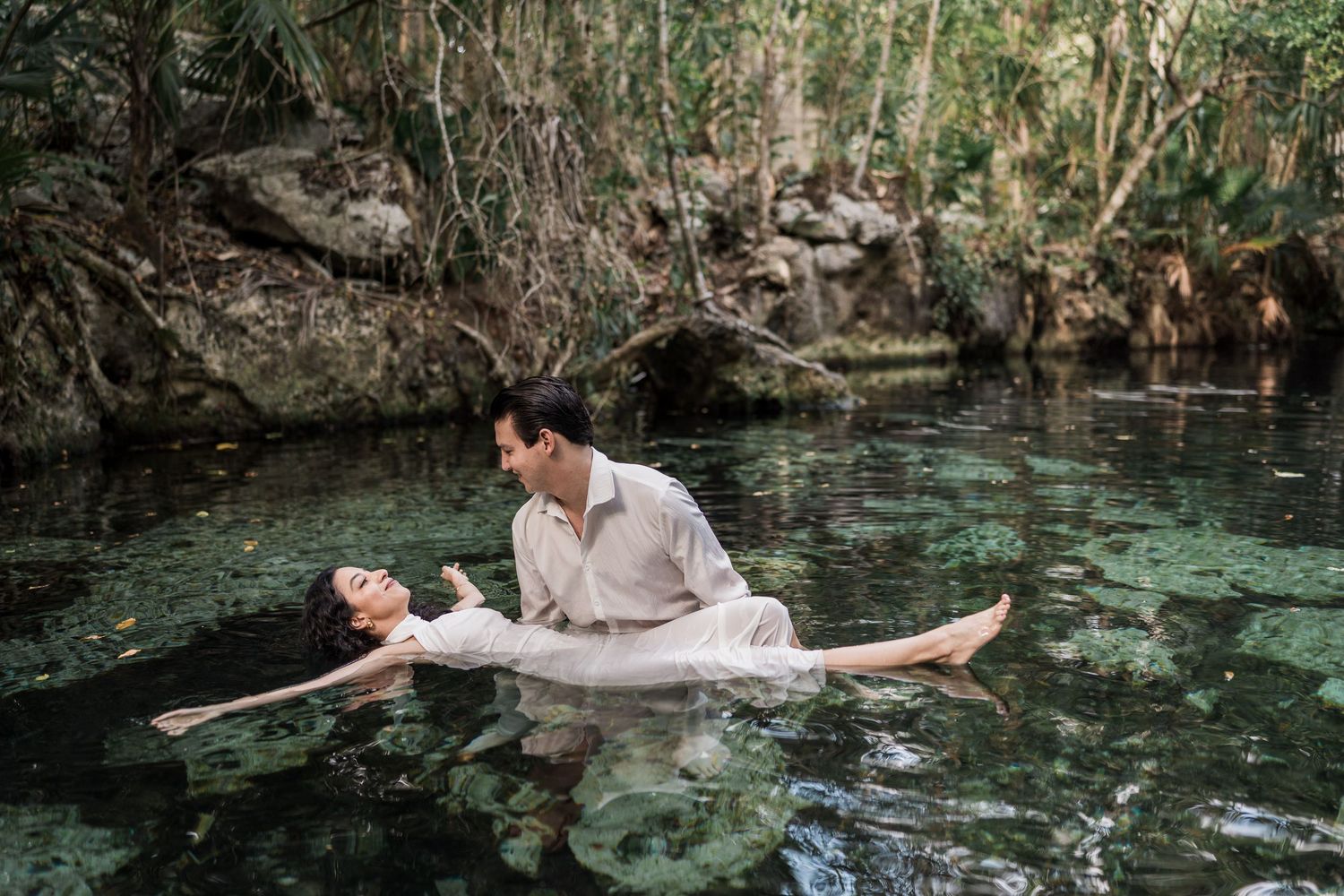 Couple in white attire posing romantically in a serene cenote surrounded by lush tropical greenery.