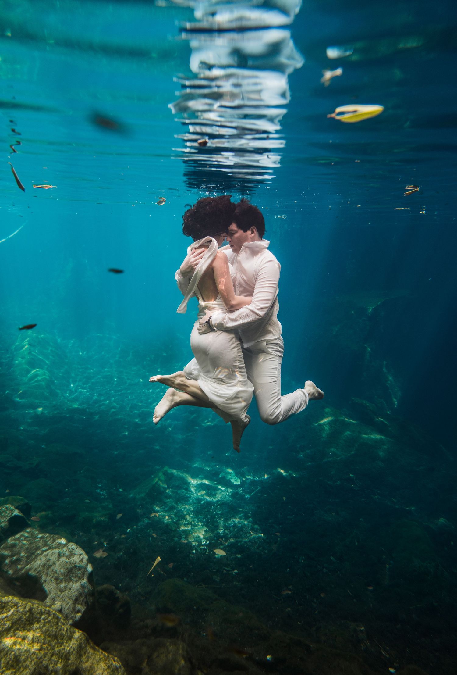 Couple in white attire sharing a romantic underwater kiss in a crystal clear blue ocean with fish swimming around them.