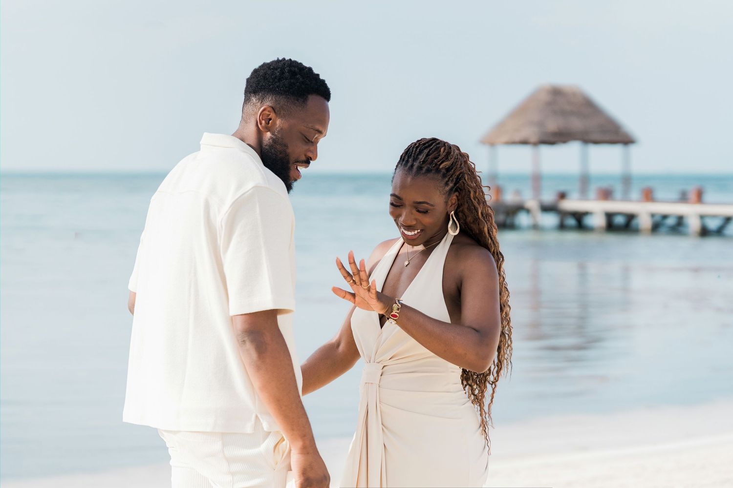 Happy couple admiring engagement ring on tropical beach near pier.