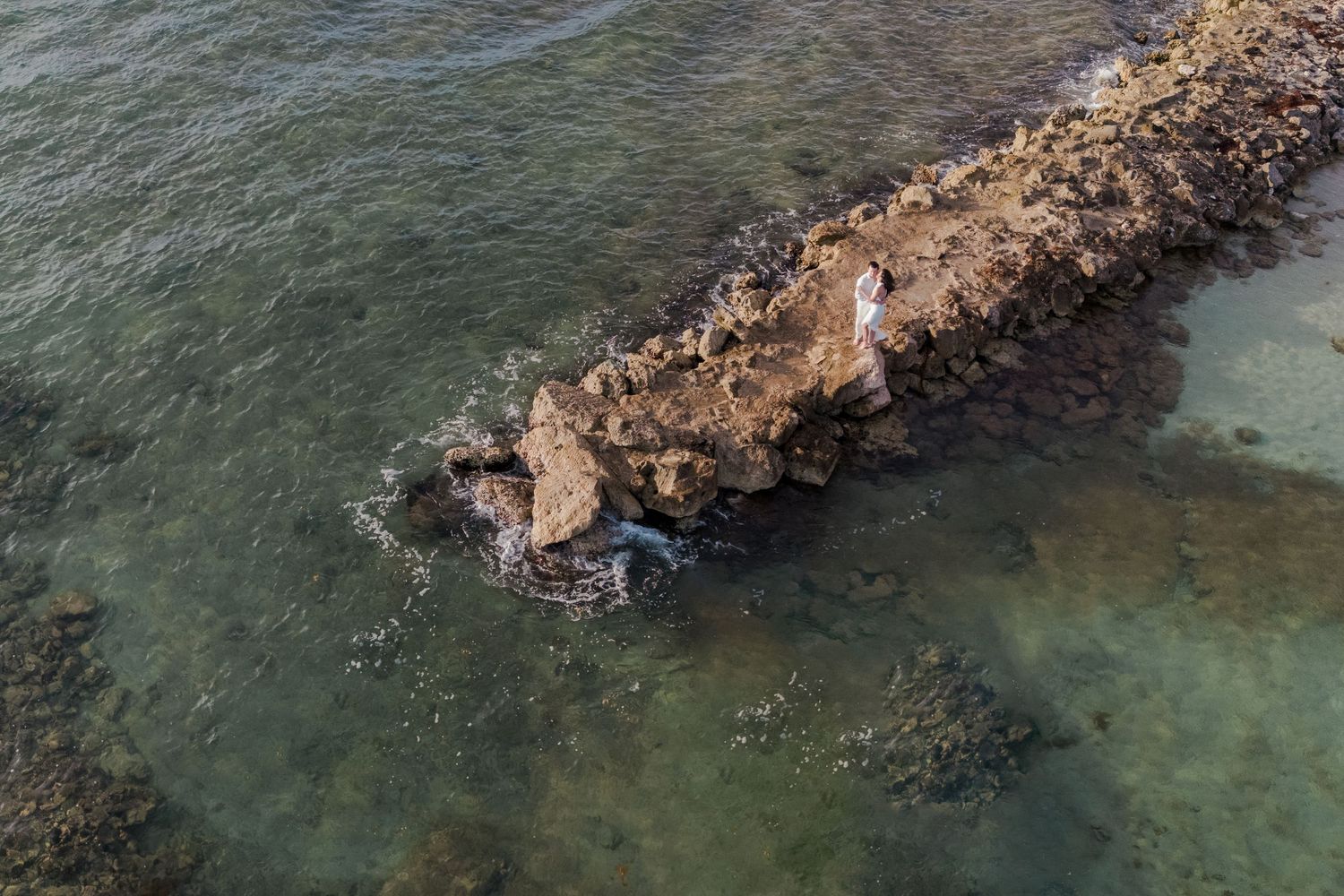 Aerial view of a rocky coastal jetty extending into clear turquoise ocean waters with a person standing on the rocks.