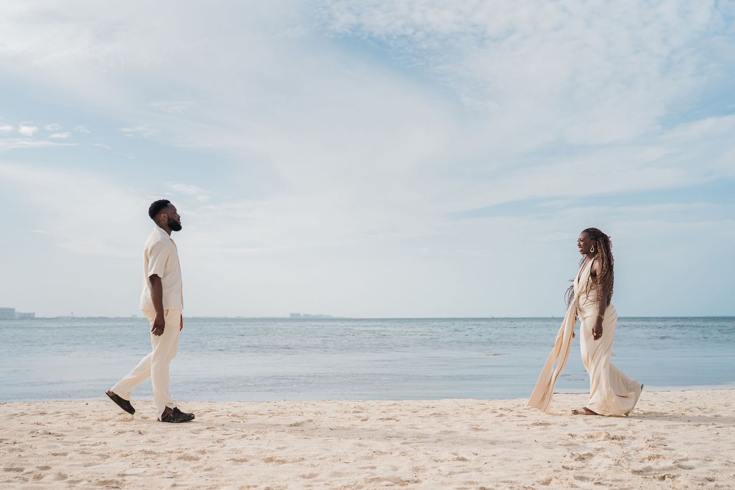A couple in white outfits walking toward each other on a sandy beach with calm ocean waters in the background.