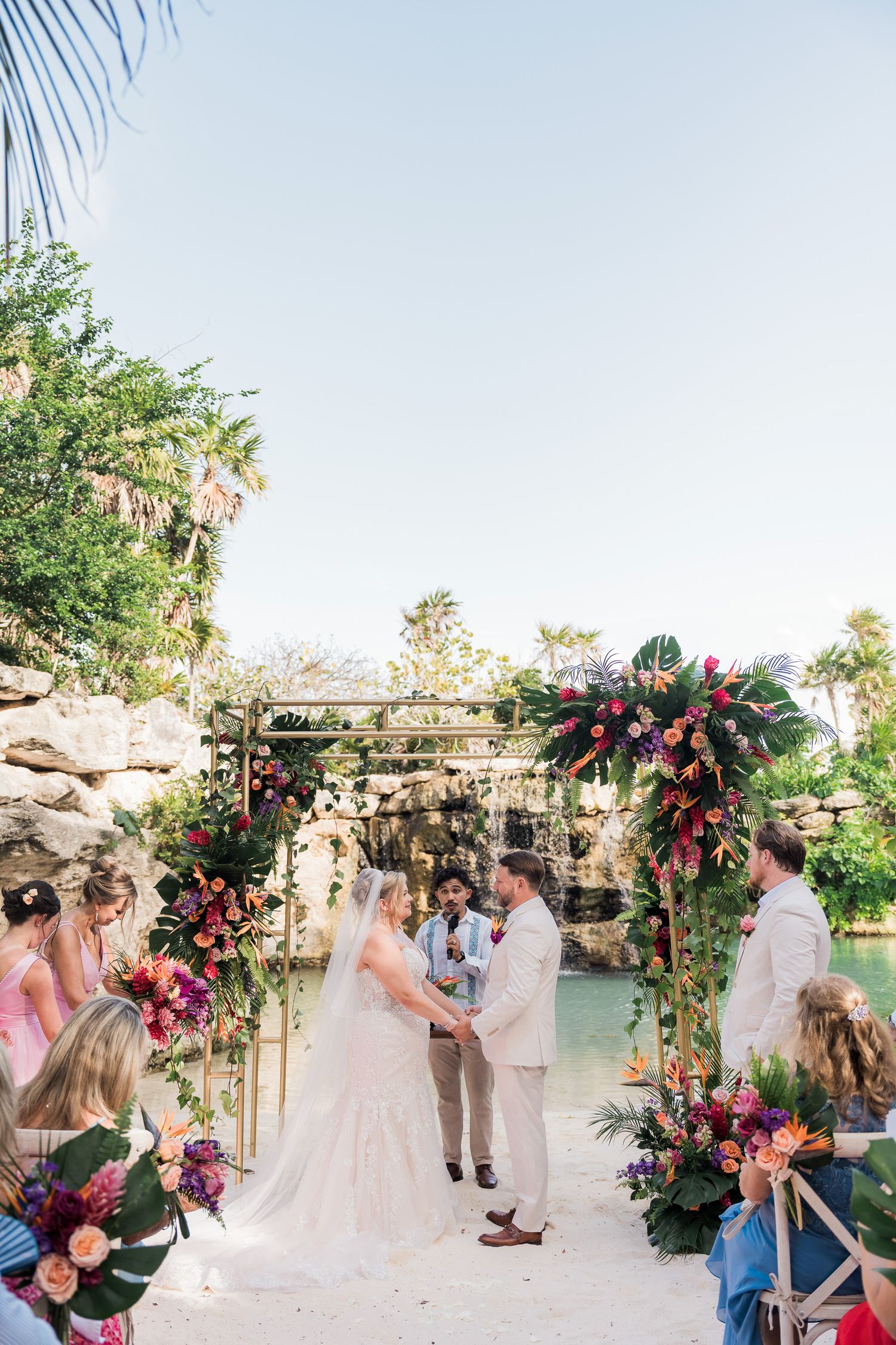 Hotel Xcaret México Wedding  - Couple exchanges vows under floral arch with tropical blooms at outdoor waterfall wedding ceremony.