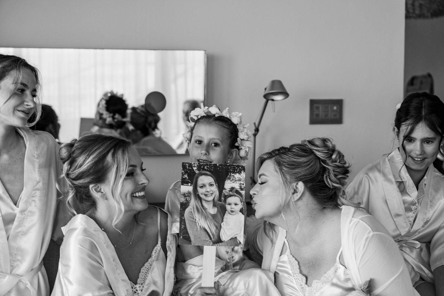 Bridesmaids in white robes laughing together while bride holds a photo during wedding preparations.