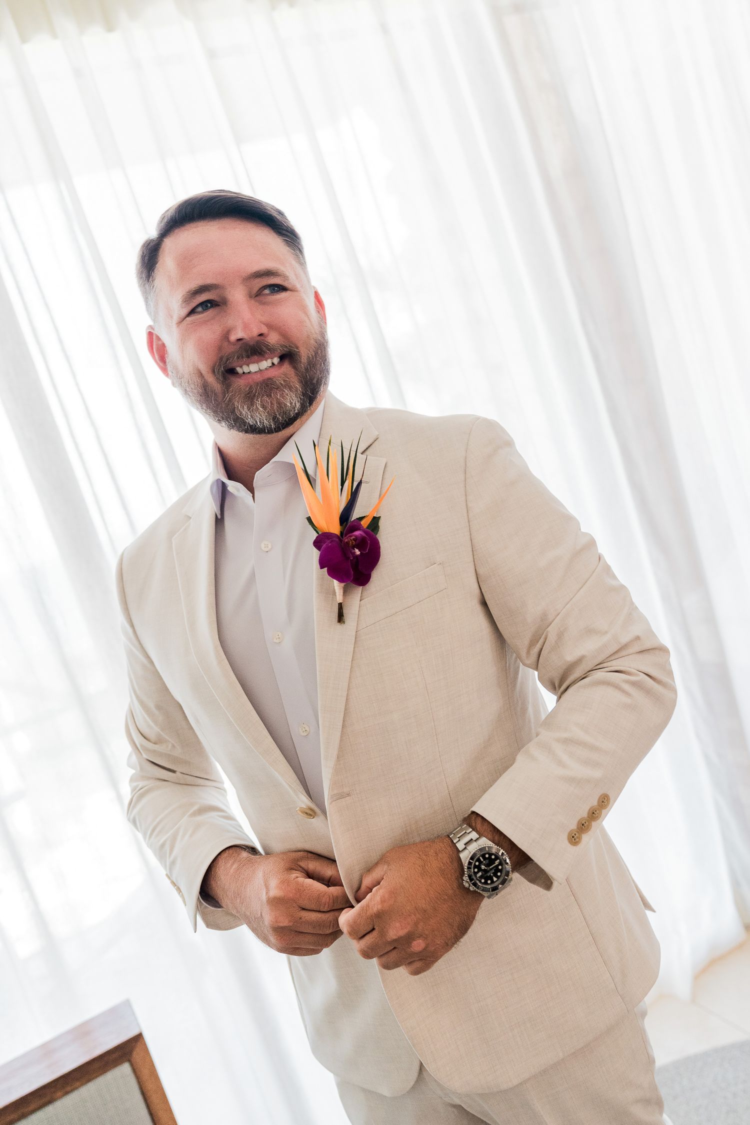 Hotel Xcaret México Wedding  - Smiling groom in beige suit with tropical boutonniere adjusting jacket button near white curtains.