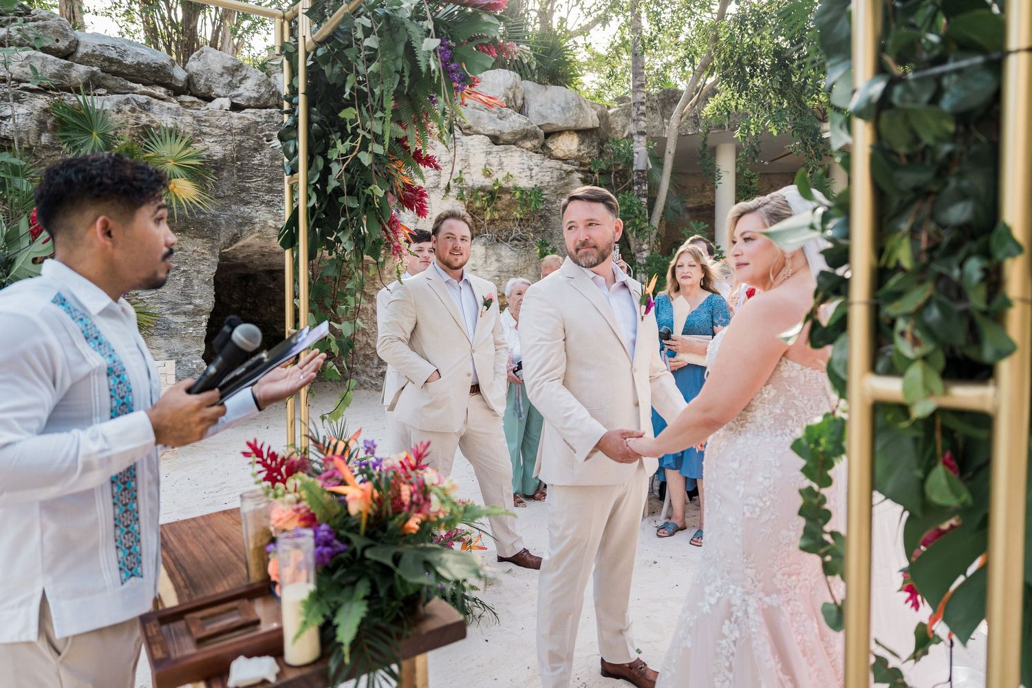 Hotel Xcaret México wedding ceremony with couple exchanging vows under floral arch, officiant reading, guests watching in tropical setting.