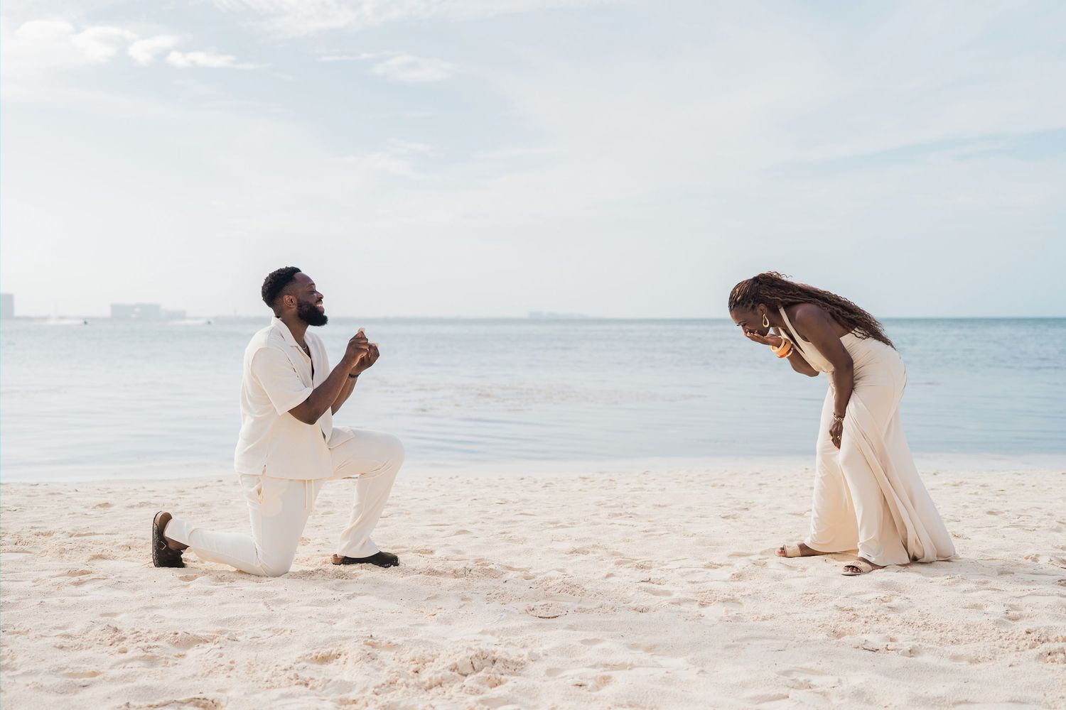Man proposes on one knee to woman on a sandy beach, both dressed in white, with calm ocean waters in the background.