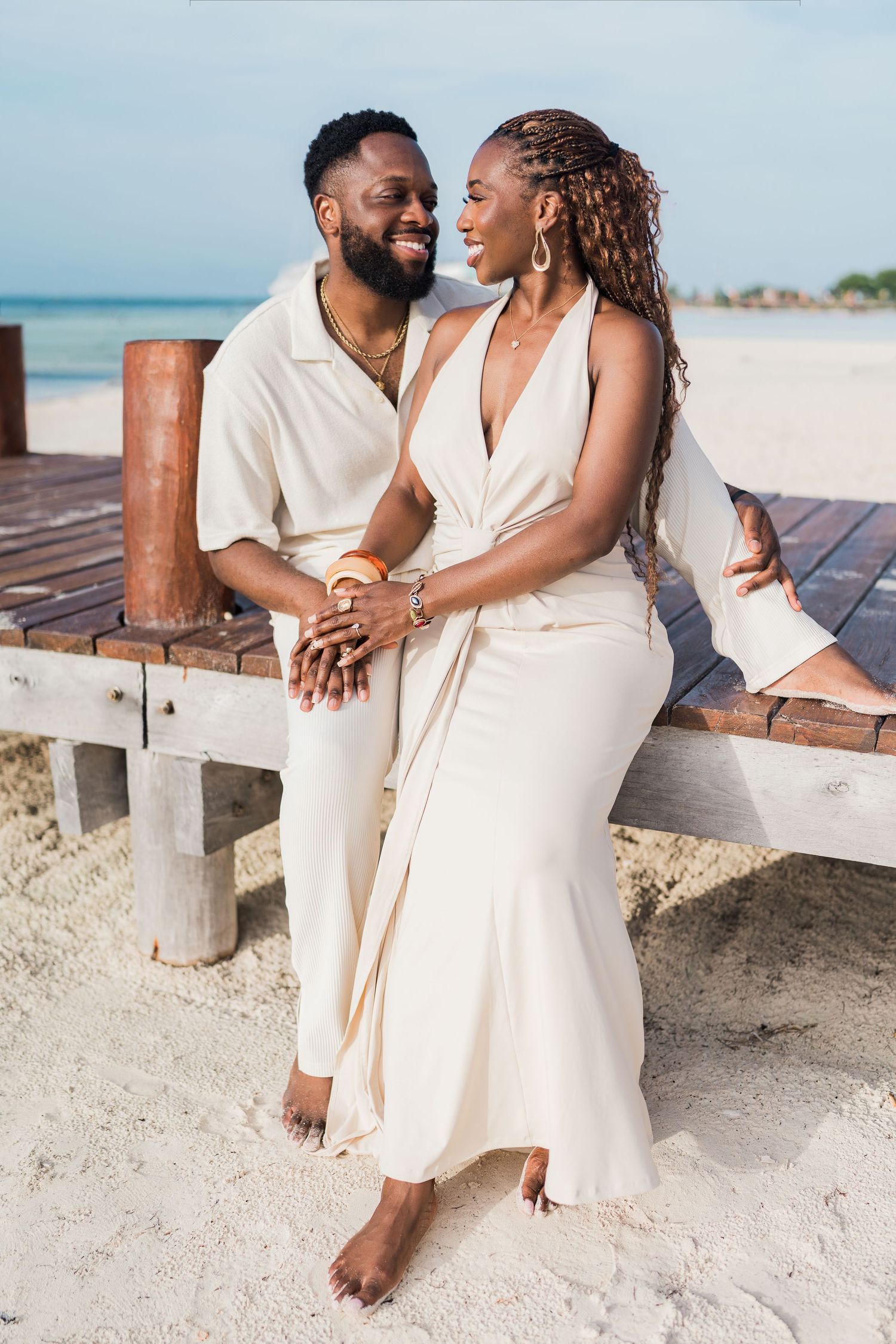 A smiling Black couple in elegant cream outfits sit closely together on a wooden beach dock, gazing at each other.