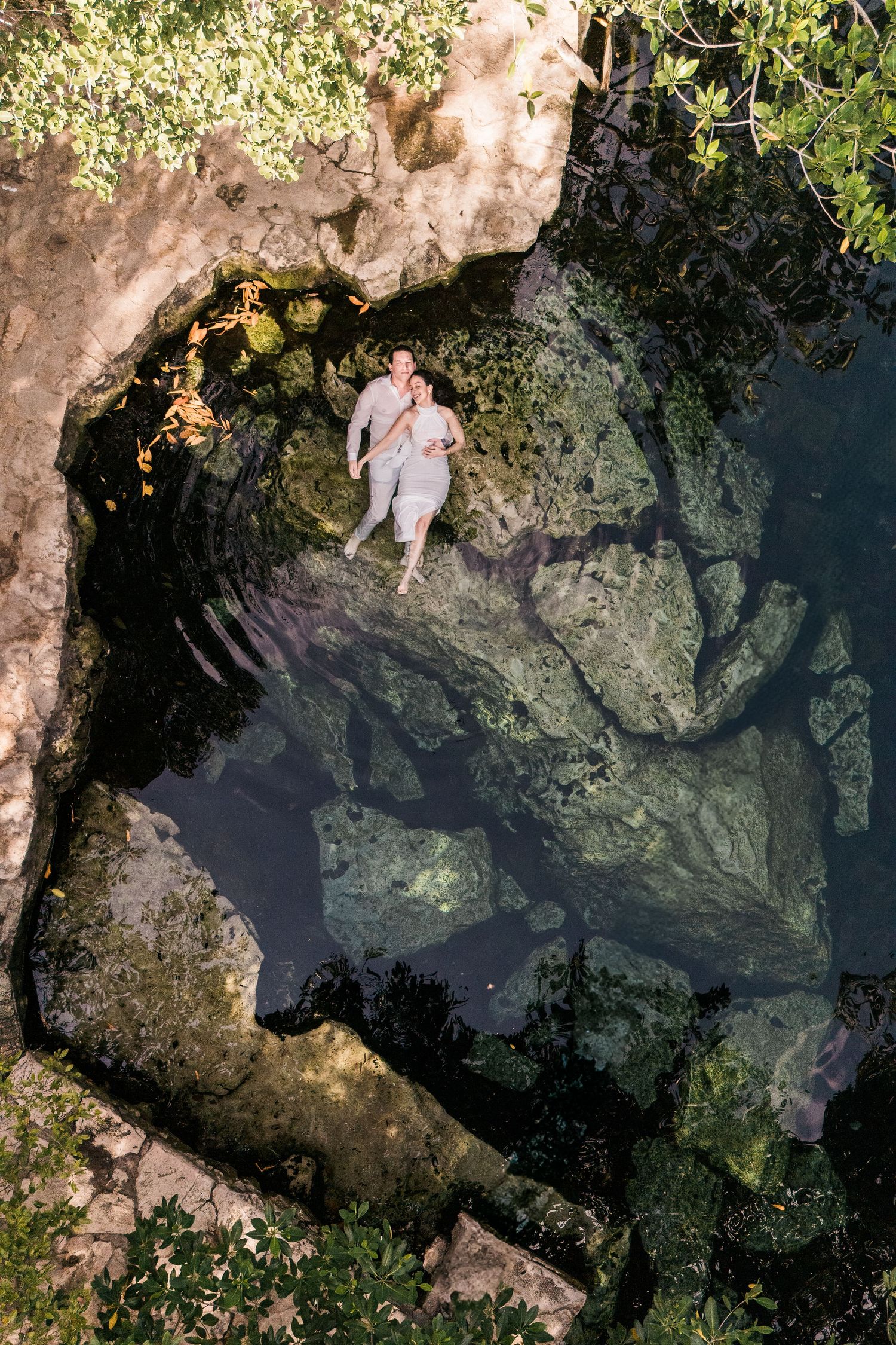 Aerial view of a couple floating together in a serene cenote with clear water and rocky walls.