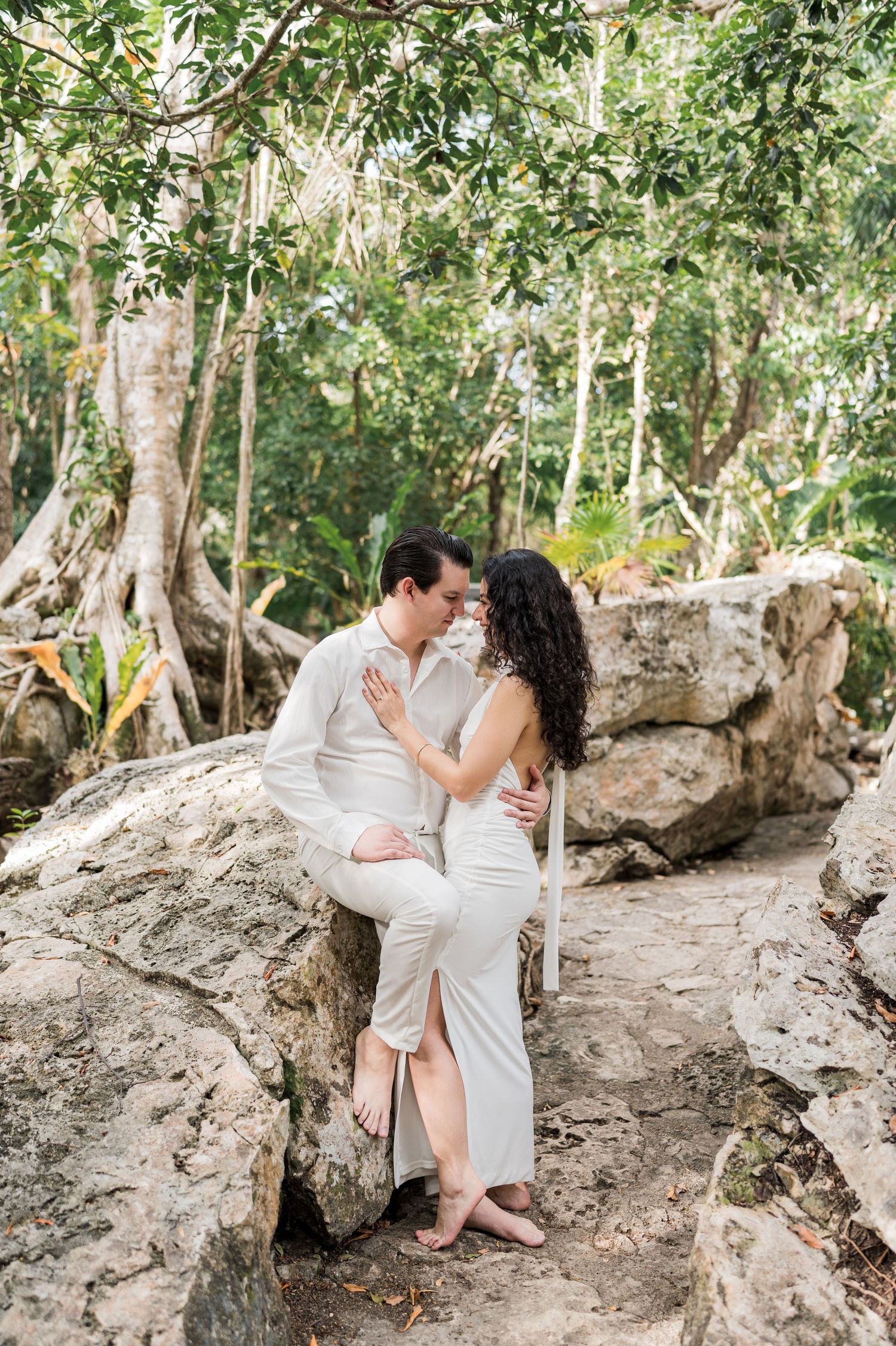 Couple in white attire sharing a romantic moment on rocks surrounded by lush tropical jungle greenery.