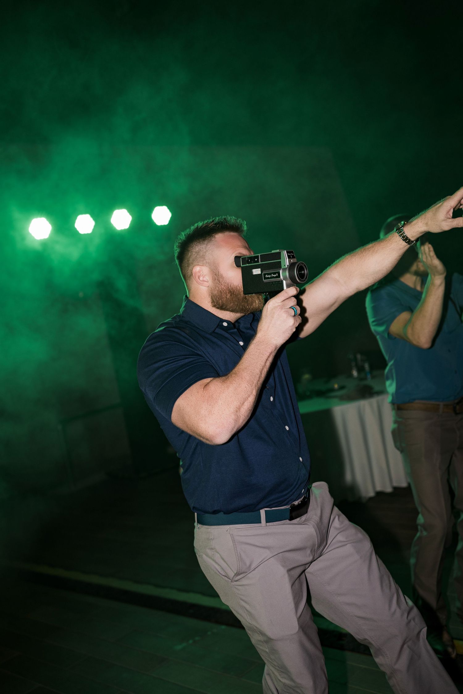 A bearded man in a navy shirt films with a vintage camera at a green-lit event venue.