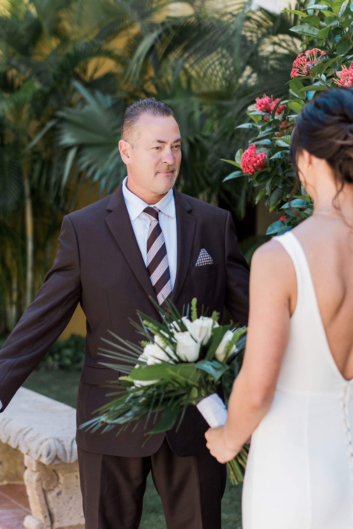 Groom in dark suit with striped tie holds bouquet during outdoor wedding ceremony surrounded by tropical greenery.
