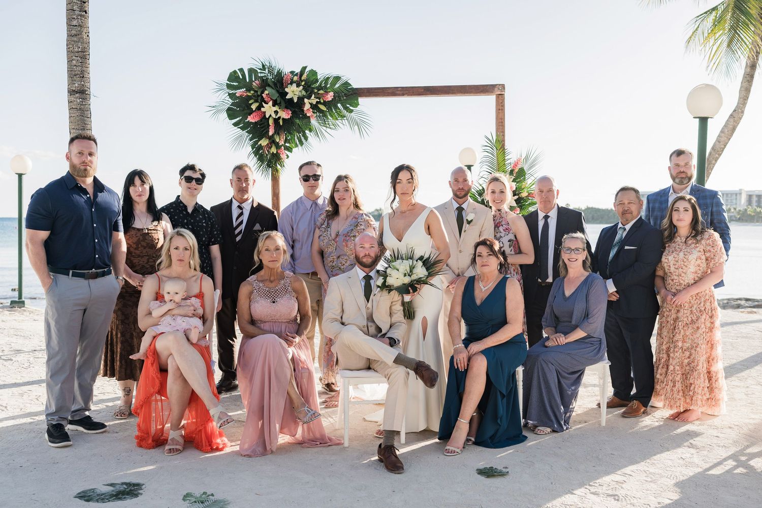 Wedding party poses on a sunny beach with a floral arch backdrop, featuring bridesmaids in pink and blue gowns.