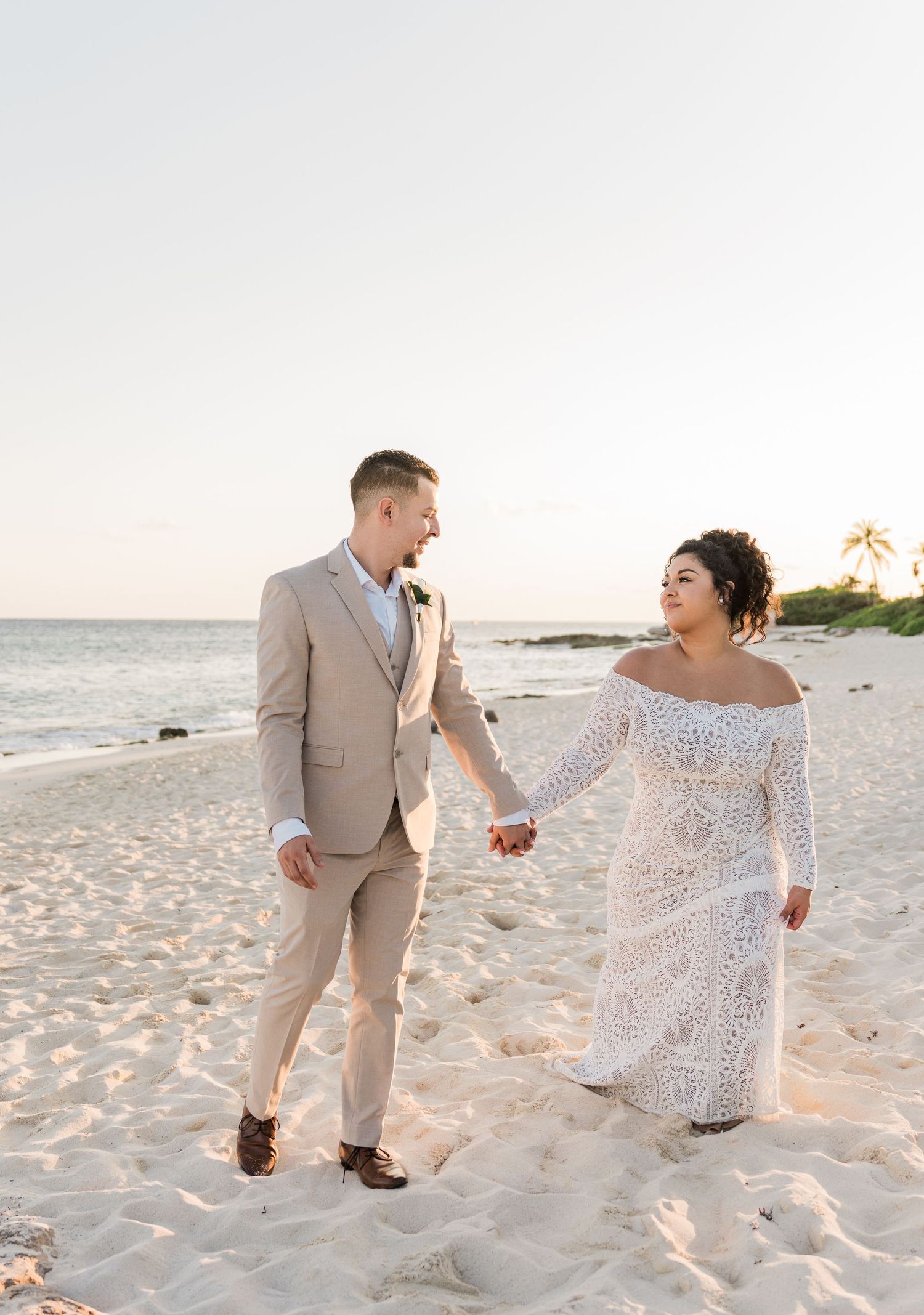 Xcaret Arte Wedding - Couple in wedding attire holding hands on a sandy beach at sunset, bride in lace gown, groom in tan suit.