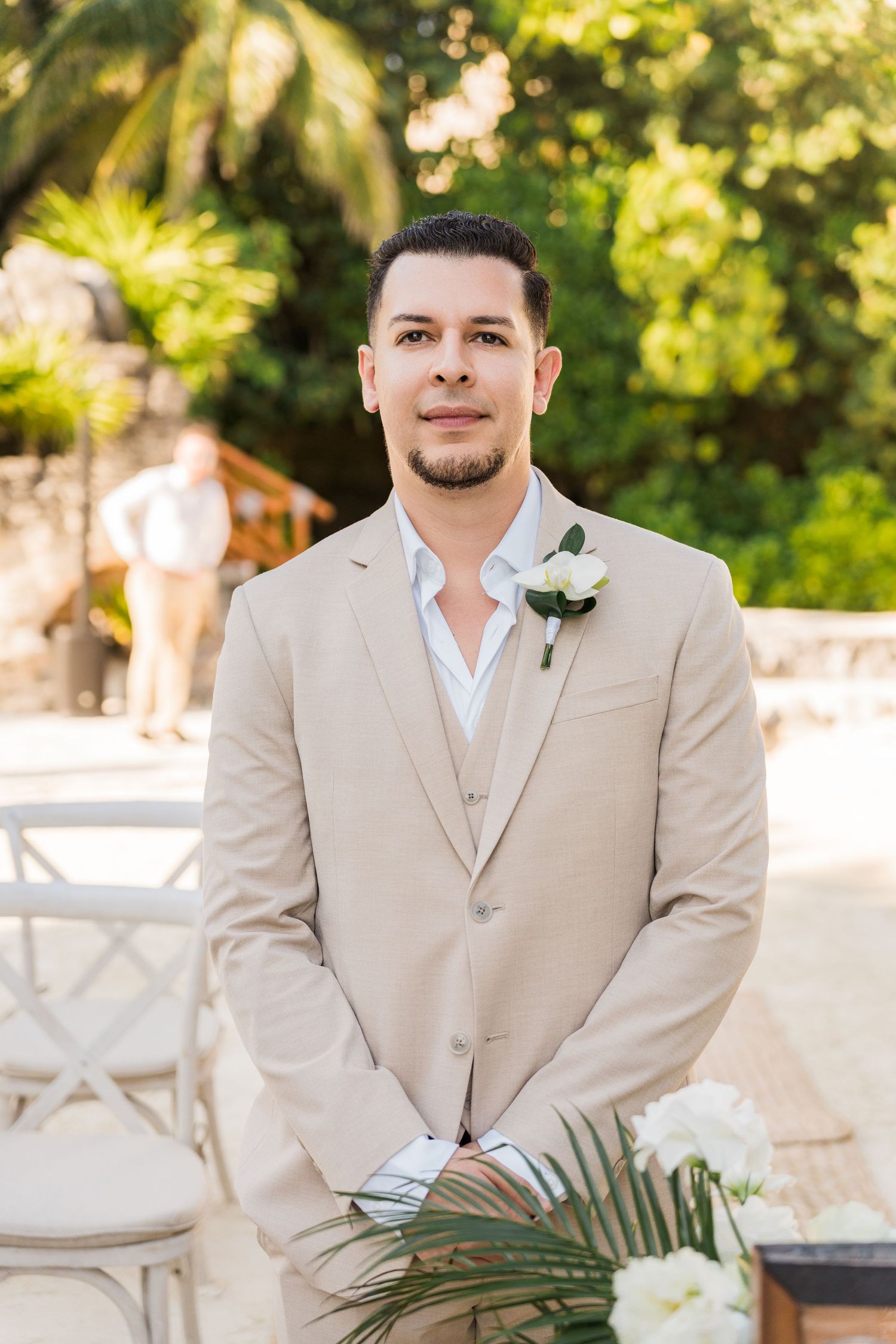 Xcaret Arte Wedding - Groom in beige suit with white boutonniere stands at outdoor wedding ceremony surrounded by tropical greenery.