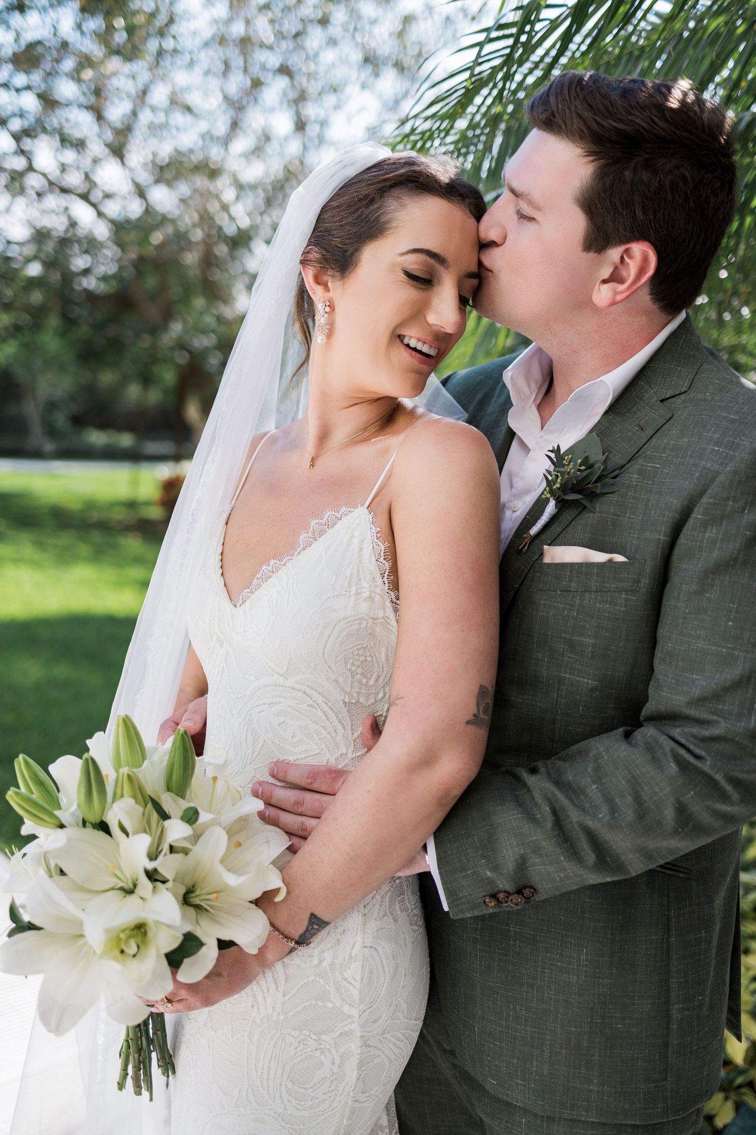 Bride in white lace gown holding lily bouquet smiles as groom in gray suit kisses her cheek outdoors.