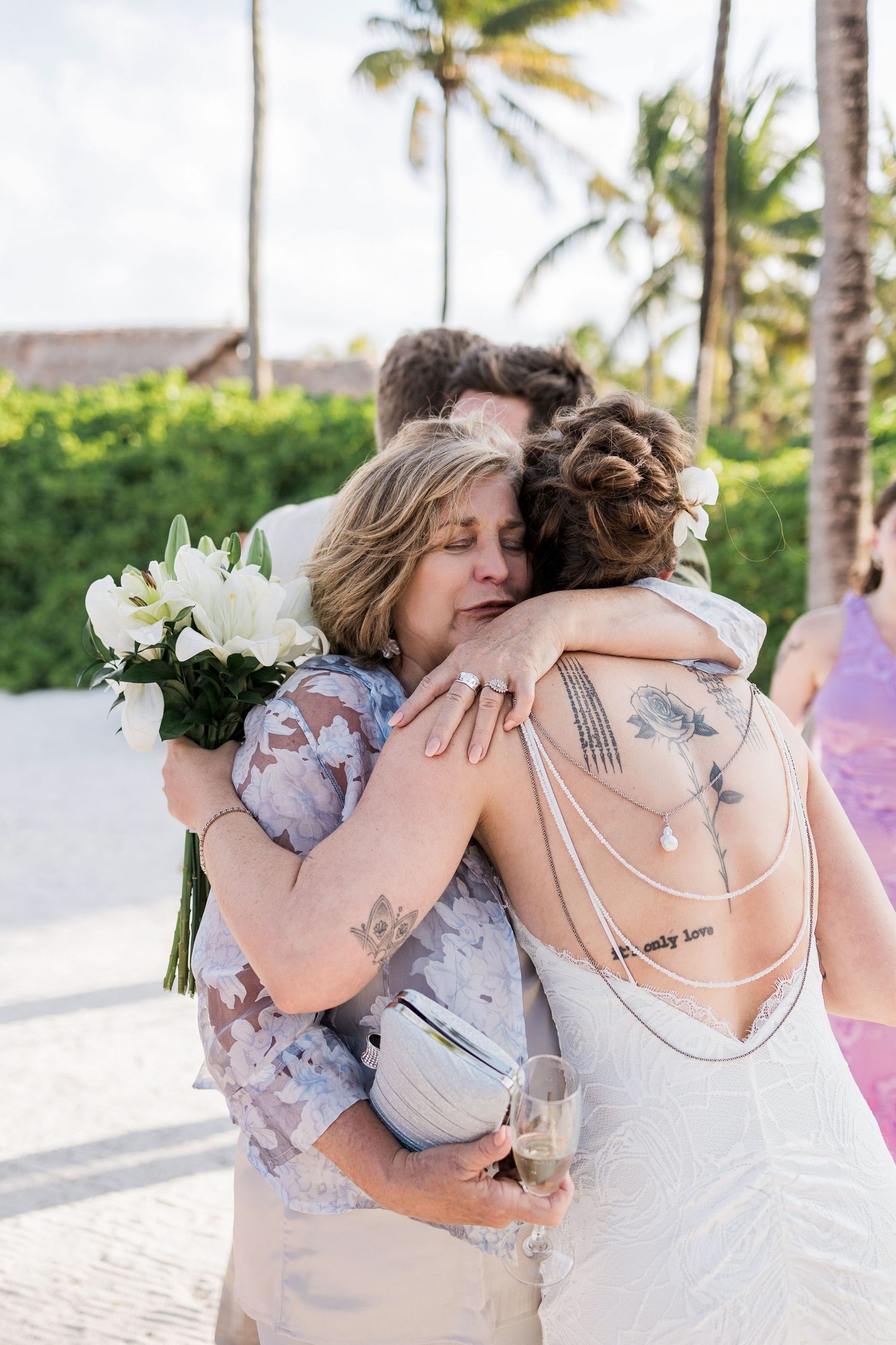 Bride with back tattoos hugs emotional guest holding white flowers at tropical beach wedding ceremony.