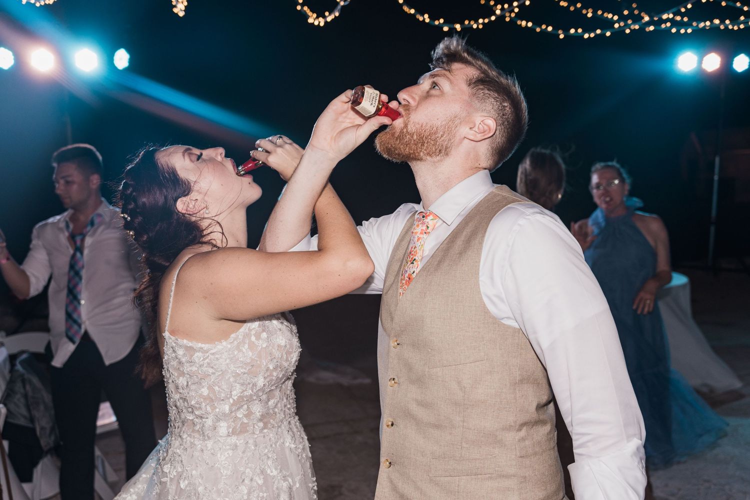 Bride feeds groom a piece of wedding cake during their lively outdoor destination wedding reception at night.