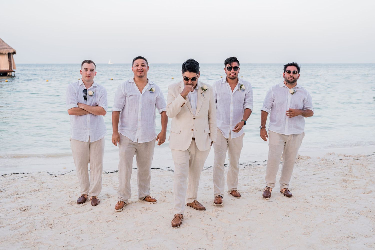 Groom in tan suit standing with groomsmen on sandy beach at dusk.
