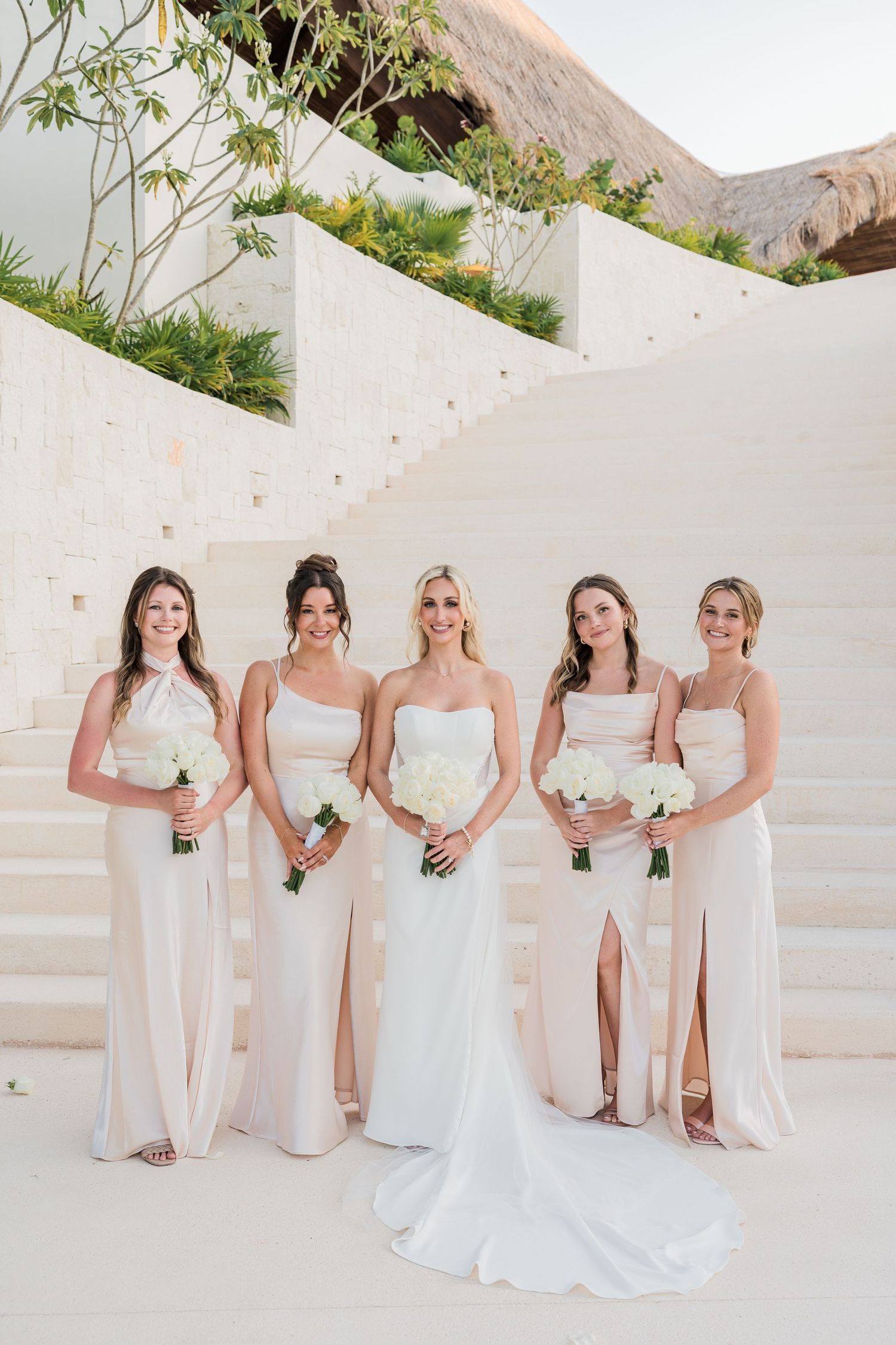 Bride and four bridesmaids in white and blush gowns holding white bouquets on elegant outdoor stairs.