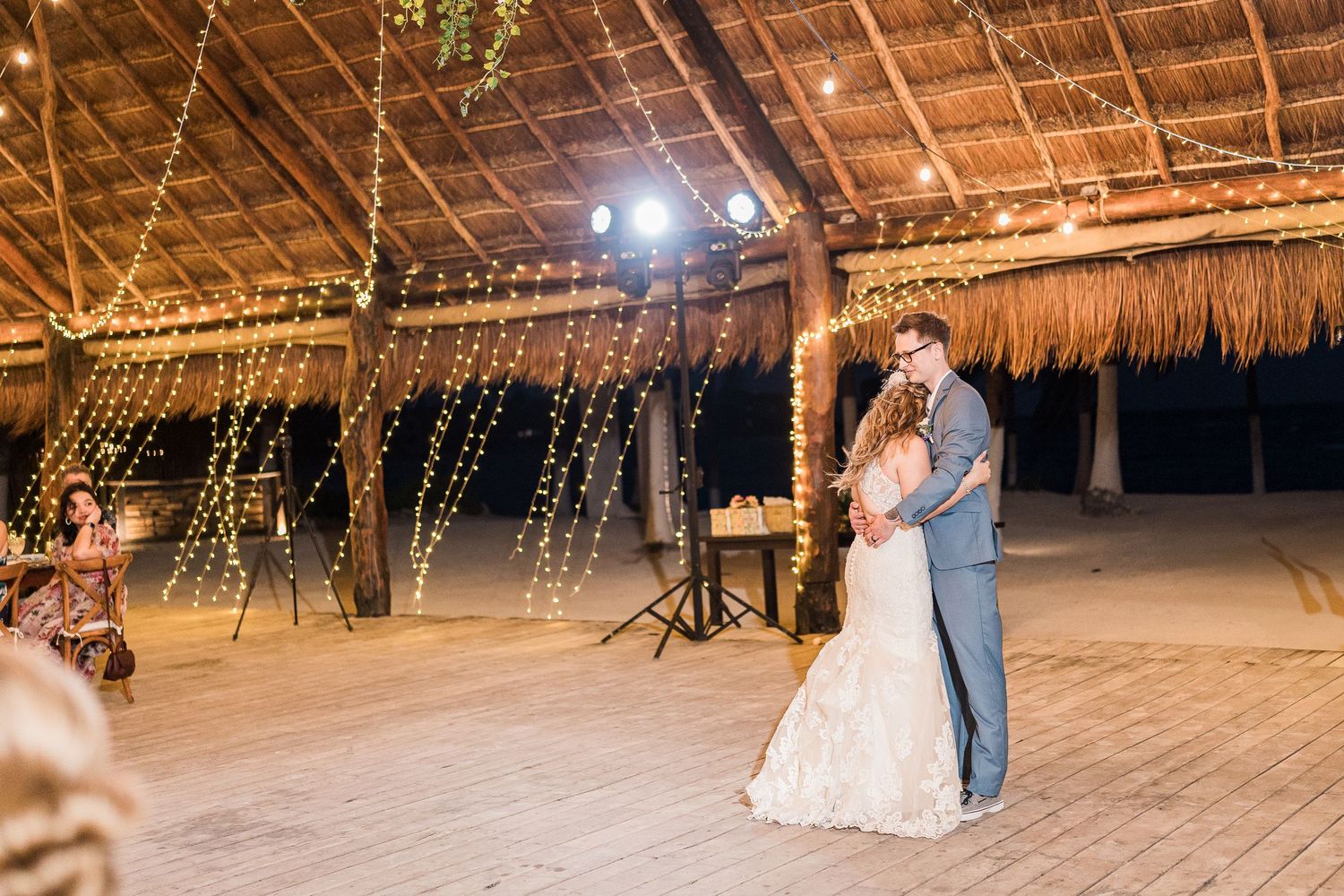 Bride and groom sharing romantic first dance under fairy lights in an open-air palapa wedding venue.
